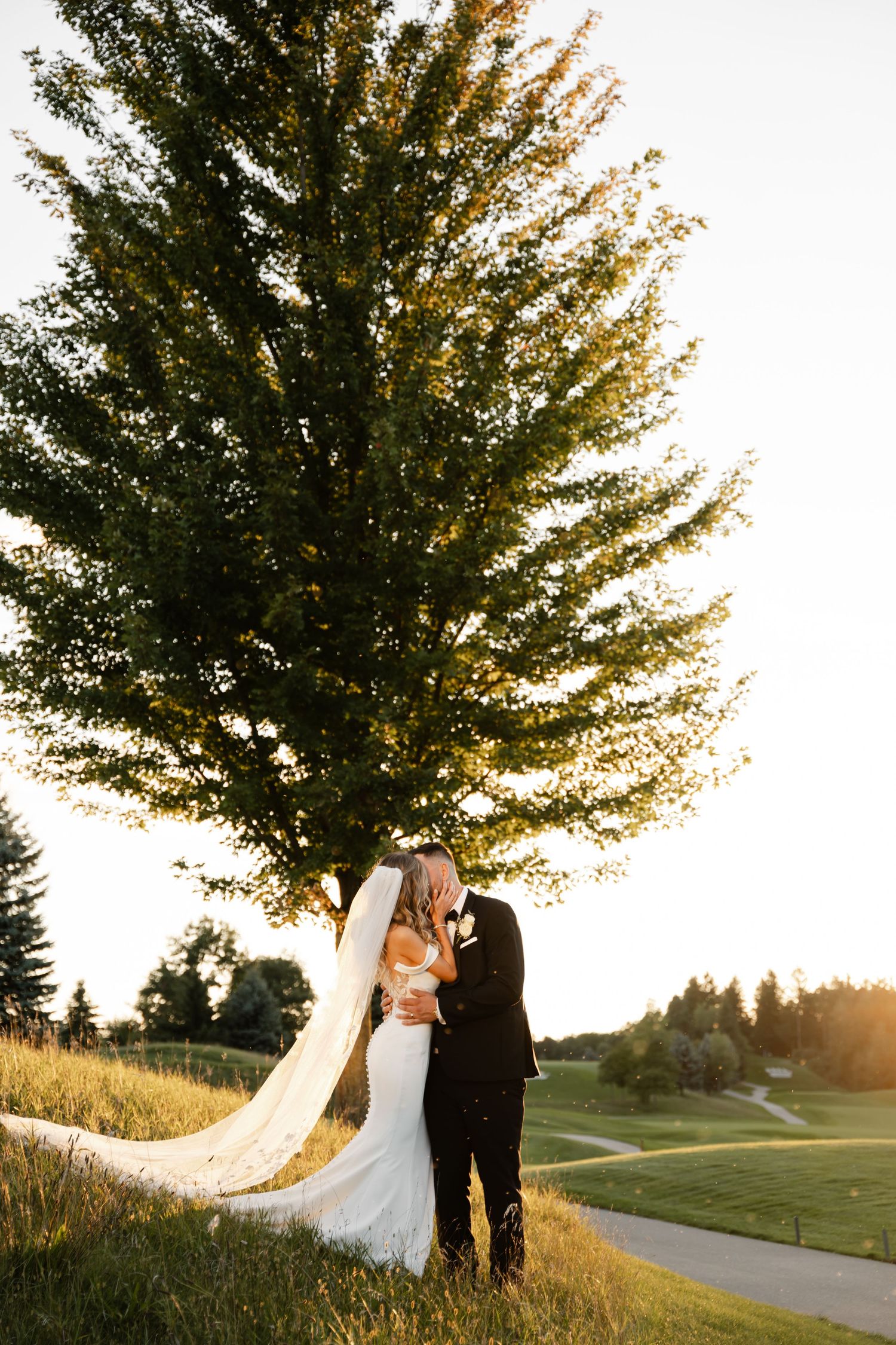A romantic wedding couple embraces under a large tree at golden hour on a golf course.