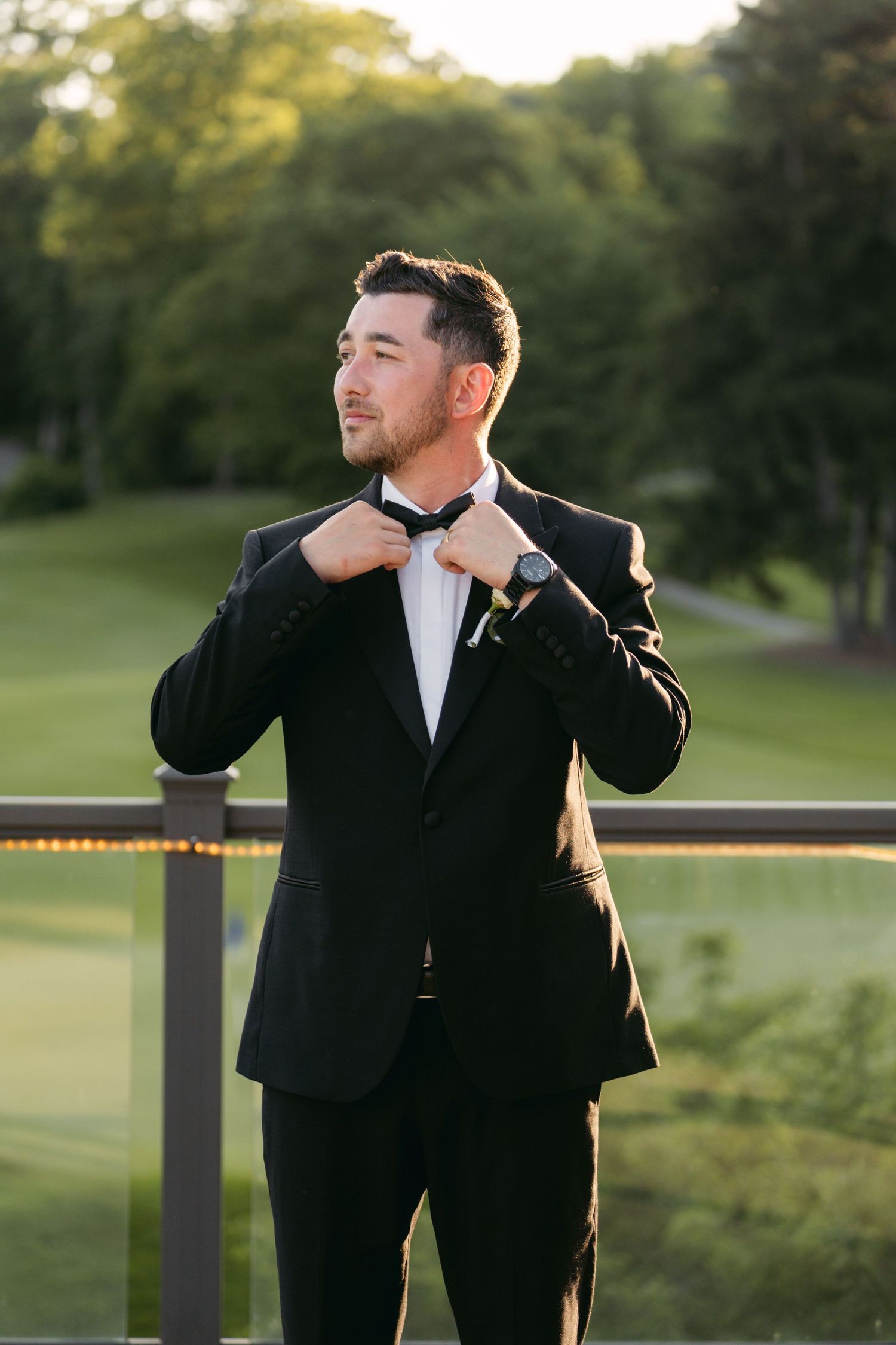Groom adjusts his bowtie while standing on an outdoor terrace at sunset.