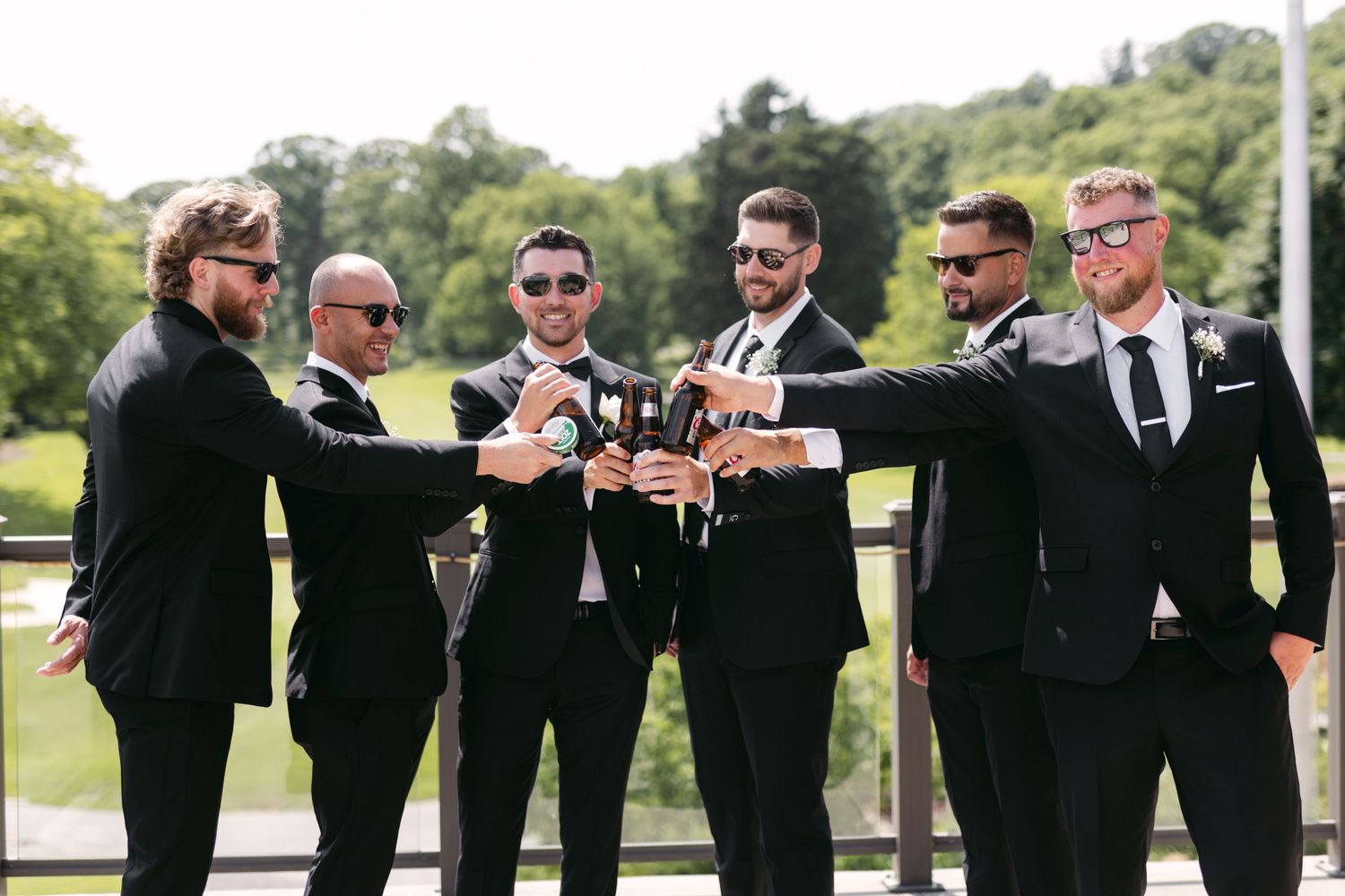 Groomsmen in black suits toast with drinks during outdoor wedding celebrations.
