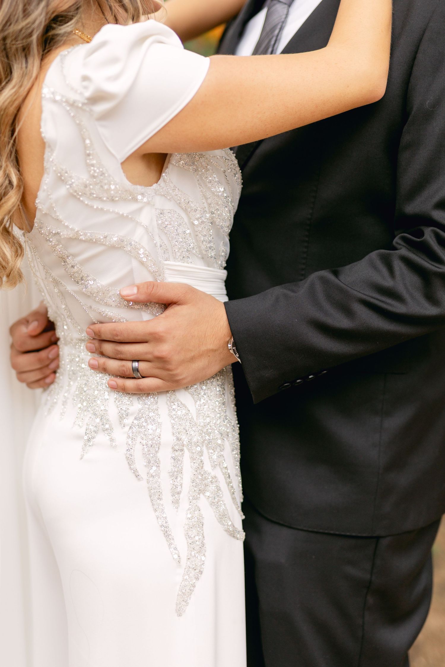 Close-up detail of sparkling white wedding dress and black tuxedo embrace.