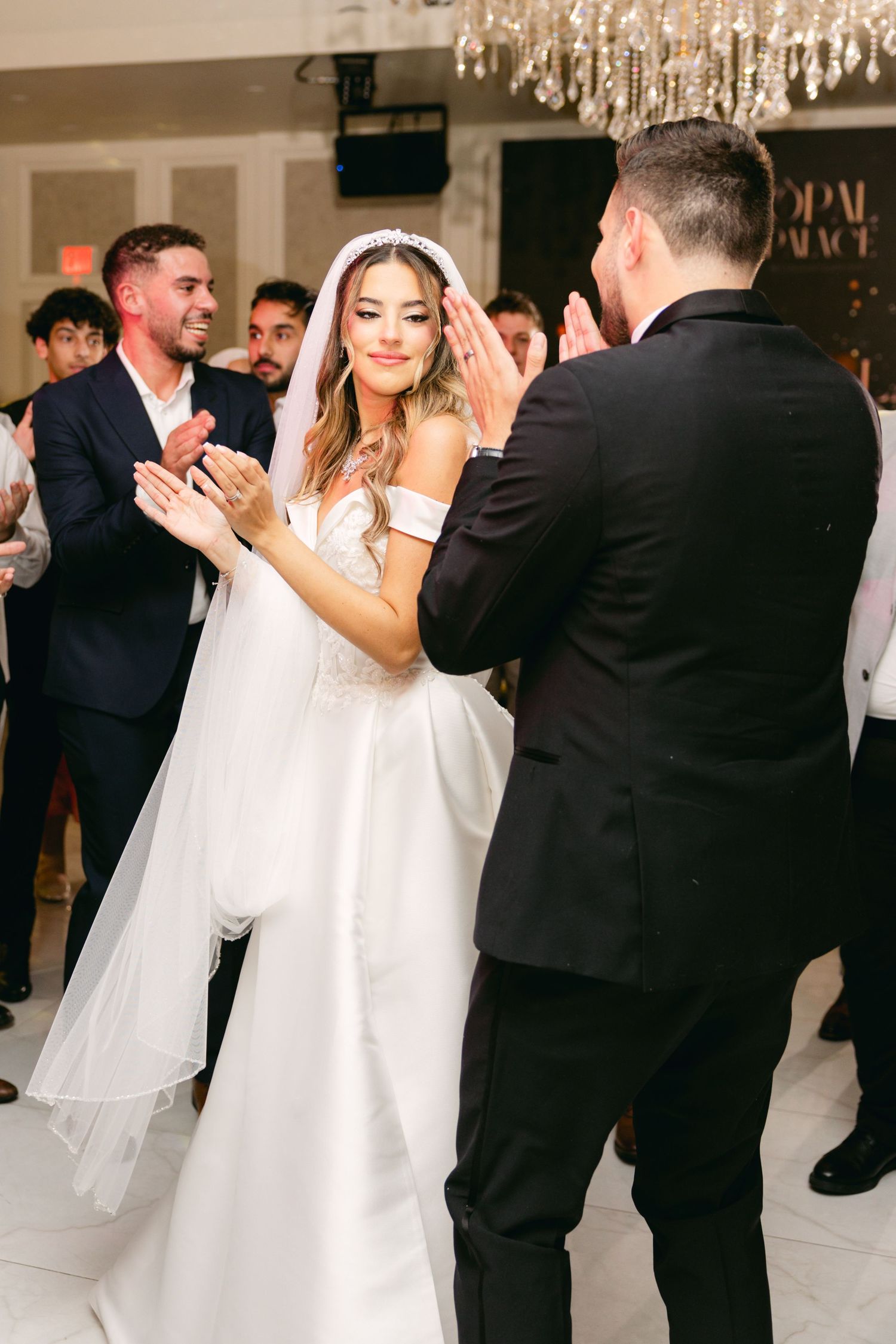 Wedding guests dancing and celebrating in an elegant white ballroom with crystal lighting.