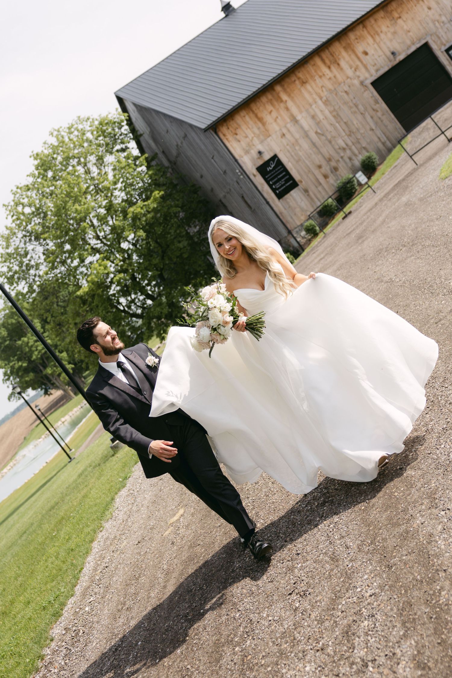 A playful wedding photo of a couple posing in front of a rustic barn with the bride's dress flowing in the wind.