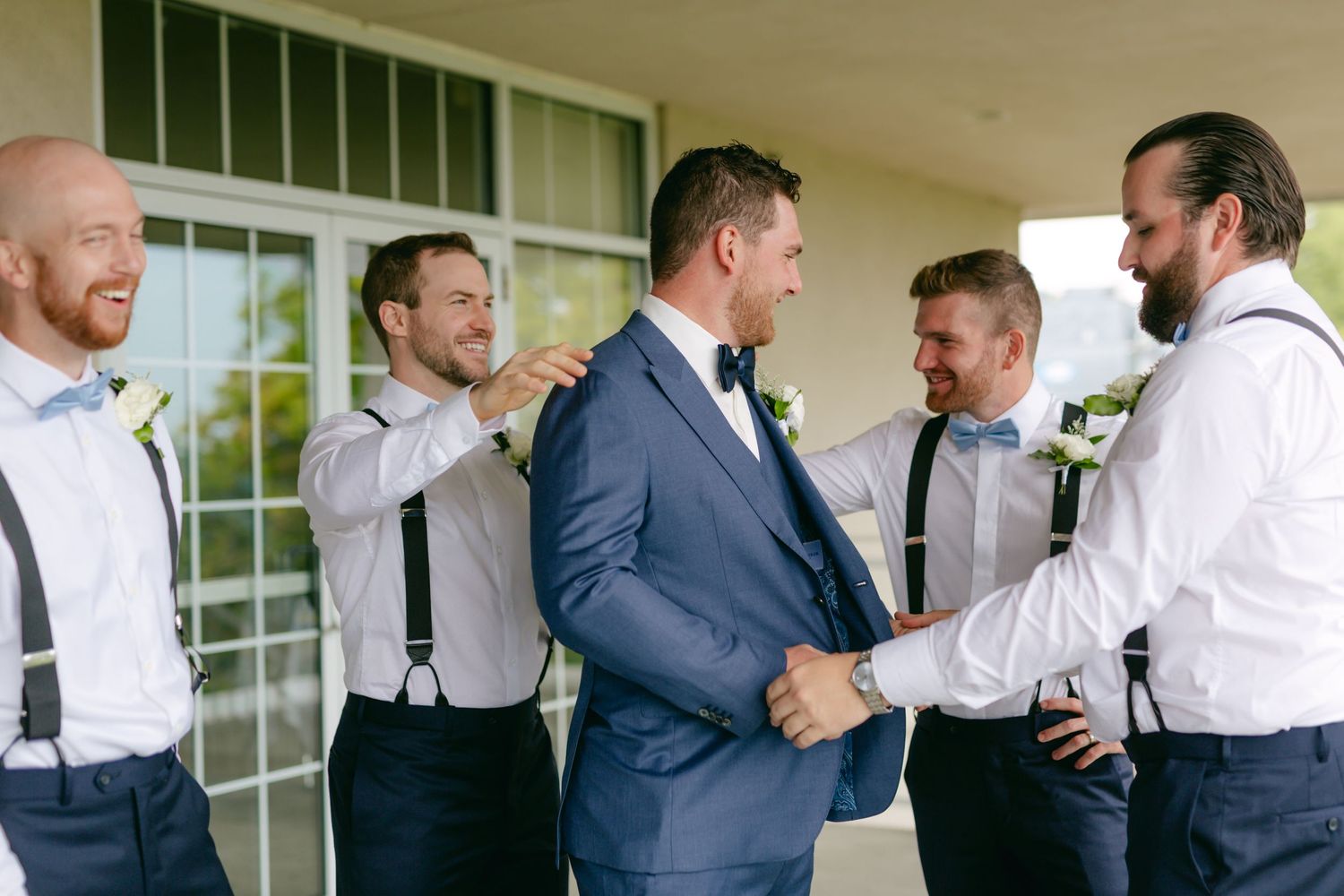 Groomsmen in white shirts and suspenders help prepare for a wedding on a covered porch.