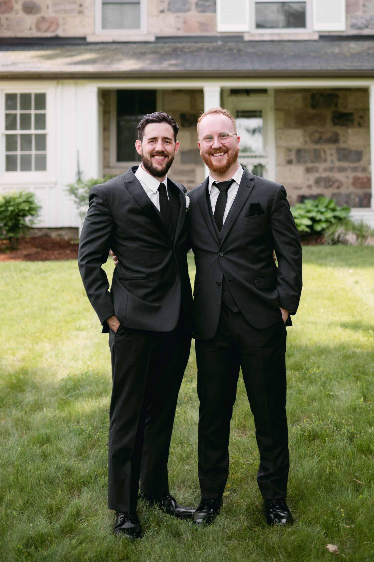 Groomsmen laugh together while posing in black suits on grass near building.