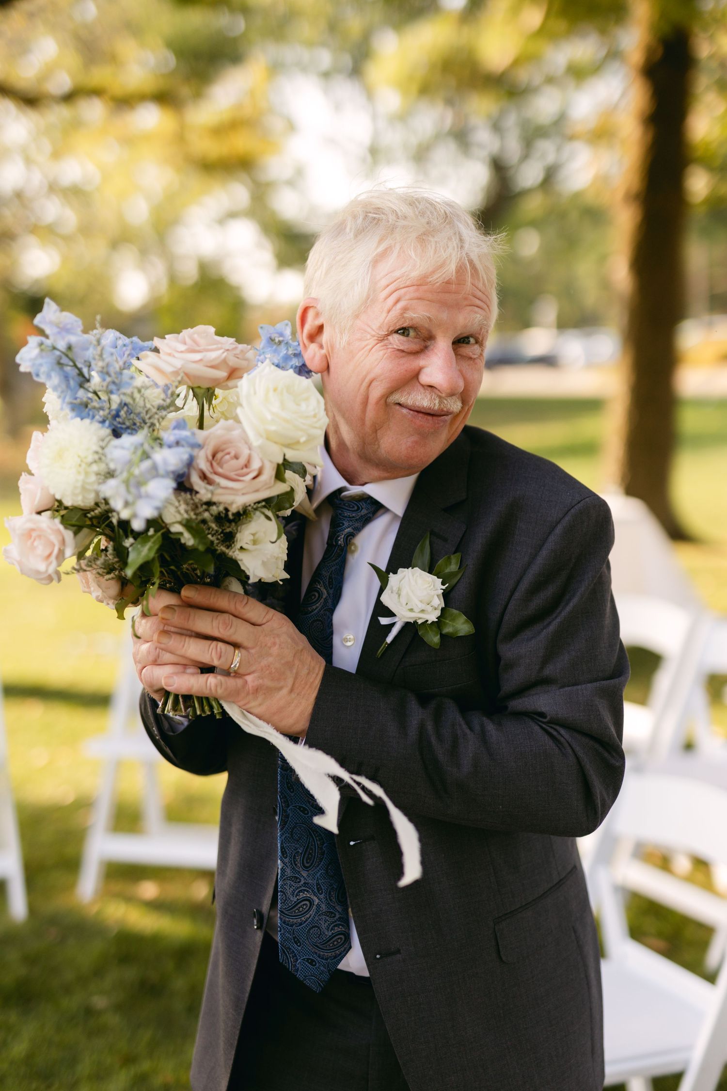 Person in dark suit holds beautiful bouquet of blue and white flowers at outdoor wedding.