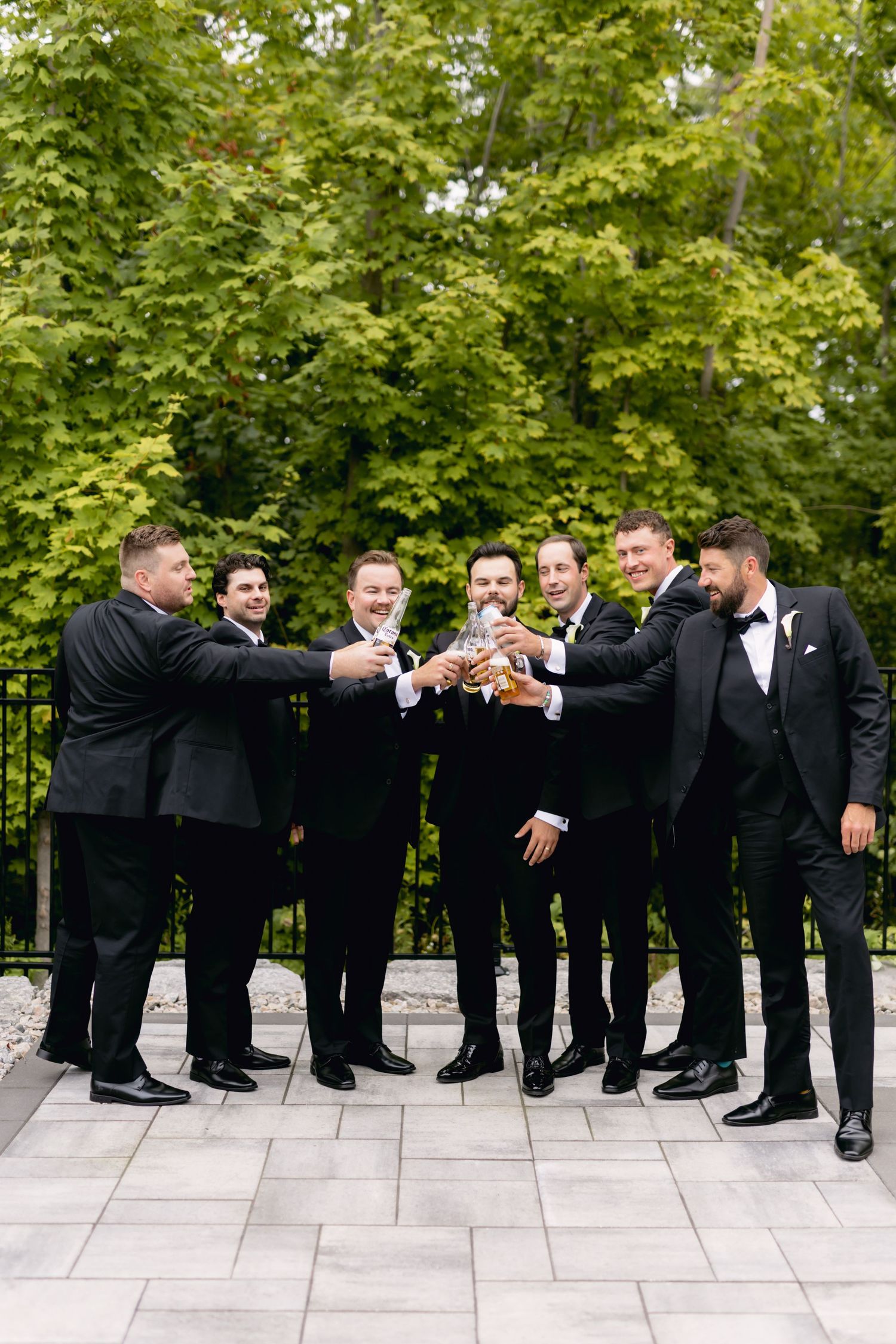 Six groomsmen in black tuxedos toast together on a stone patio surrounded by green trees at a wedding.