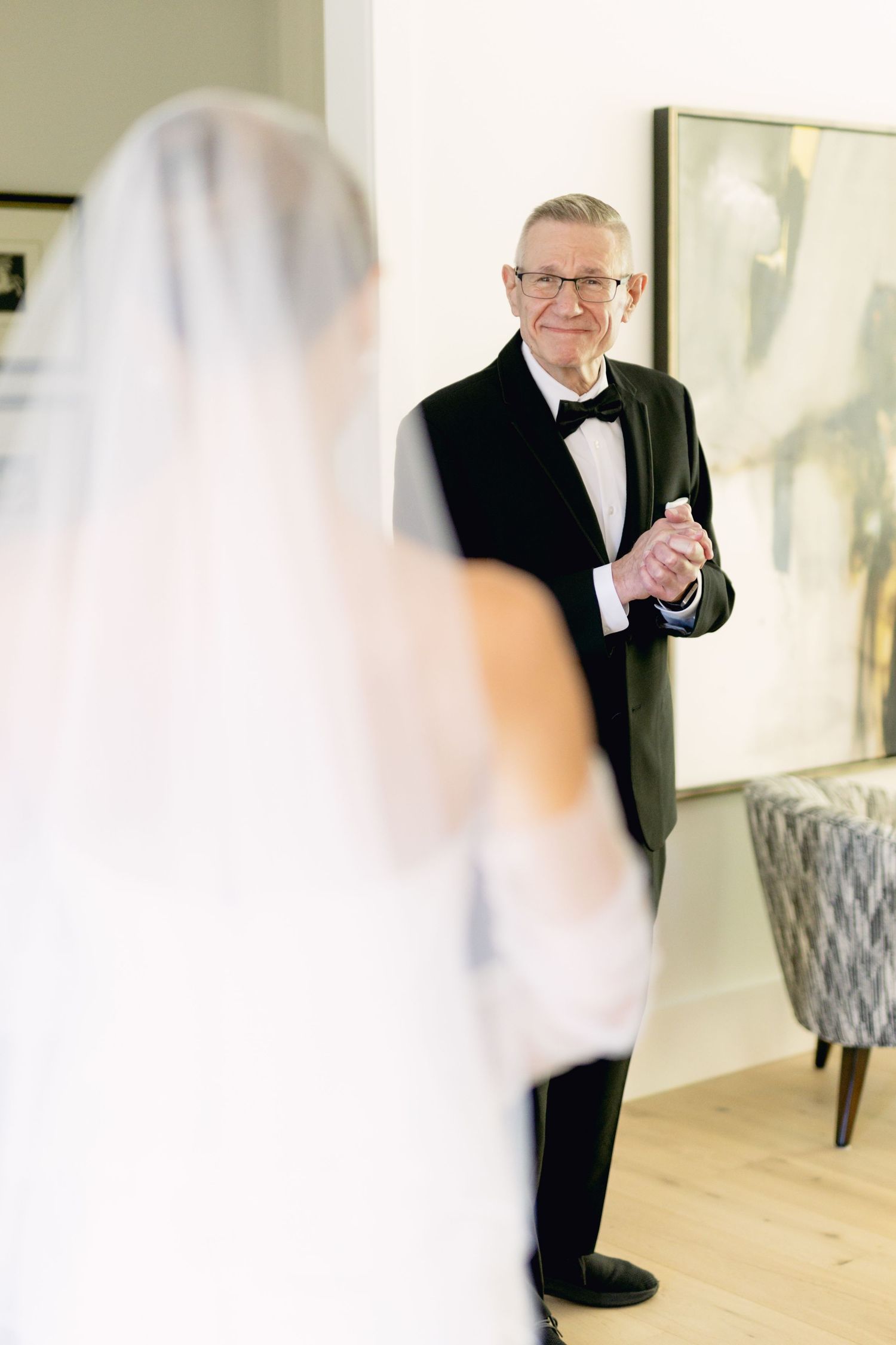 Emotional father in black tuxedo smiles at bride in white wedding gown and veil during first look moment.