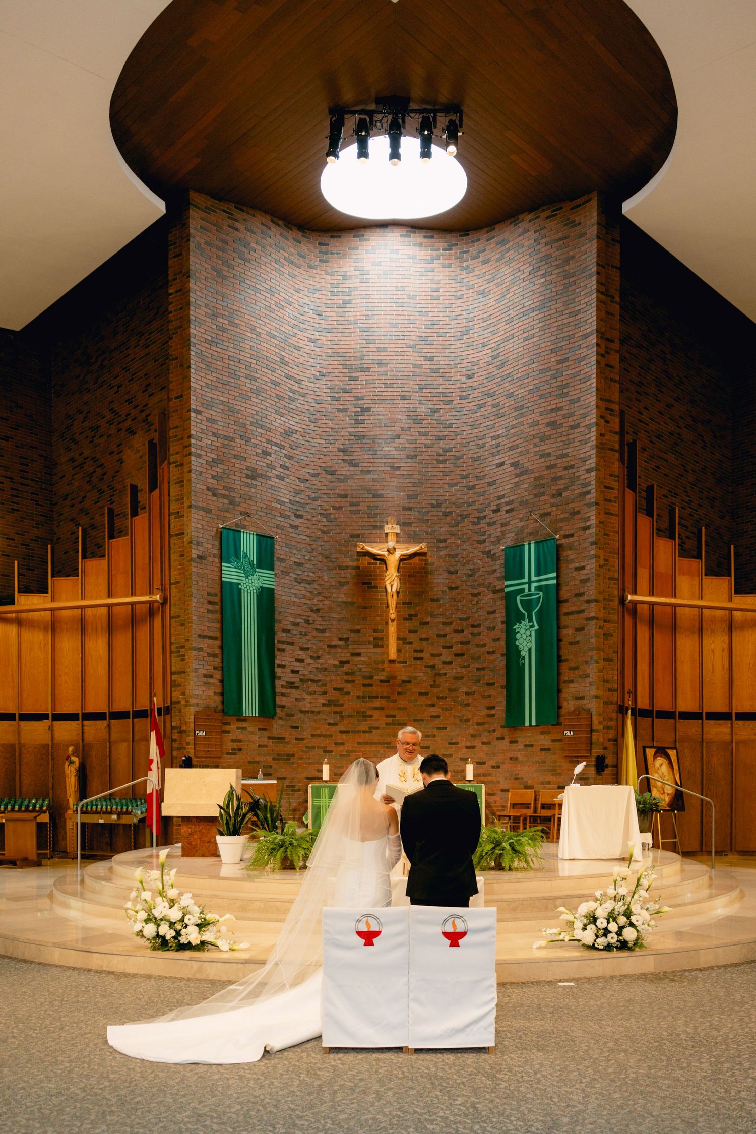 Bride and groom kneeling at church altar during Catholic wedding ceremony with floral arrangements.