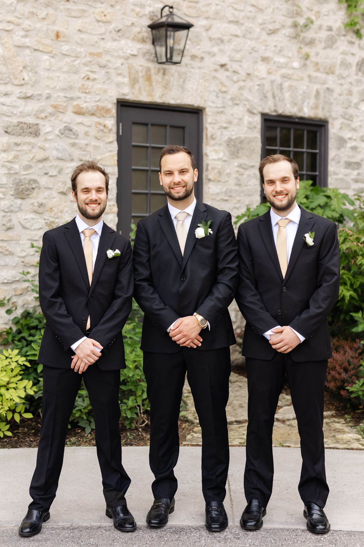 Three groomsmen in black suits with blush ties and boutonnieres pose outside a stone building at a wedding.