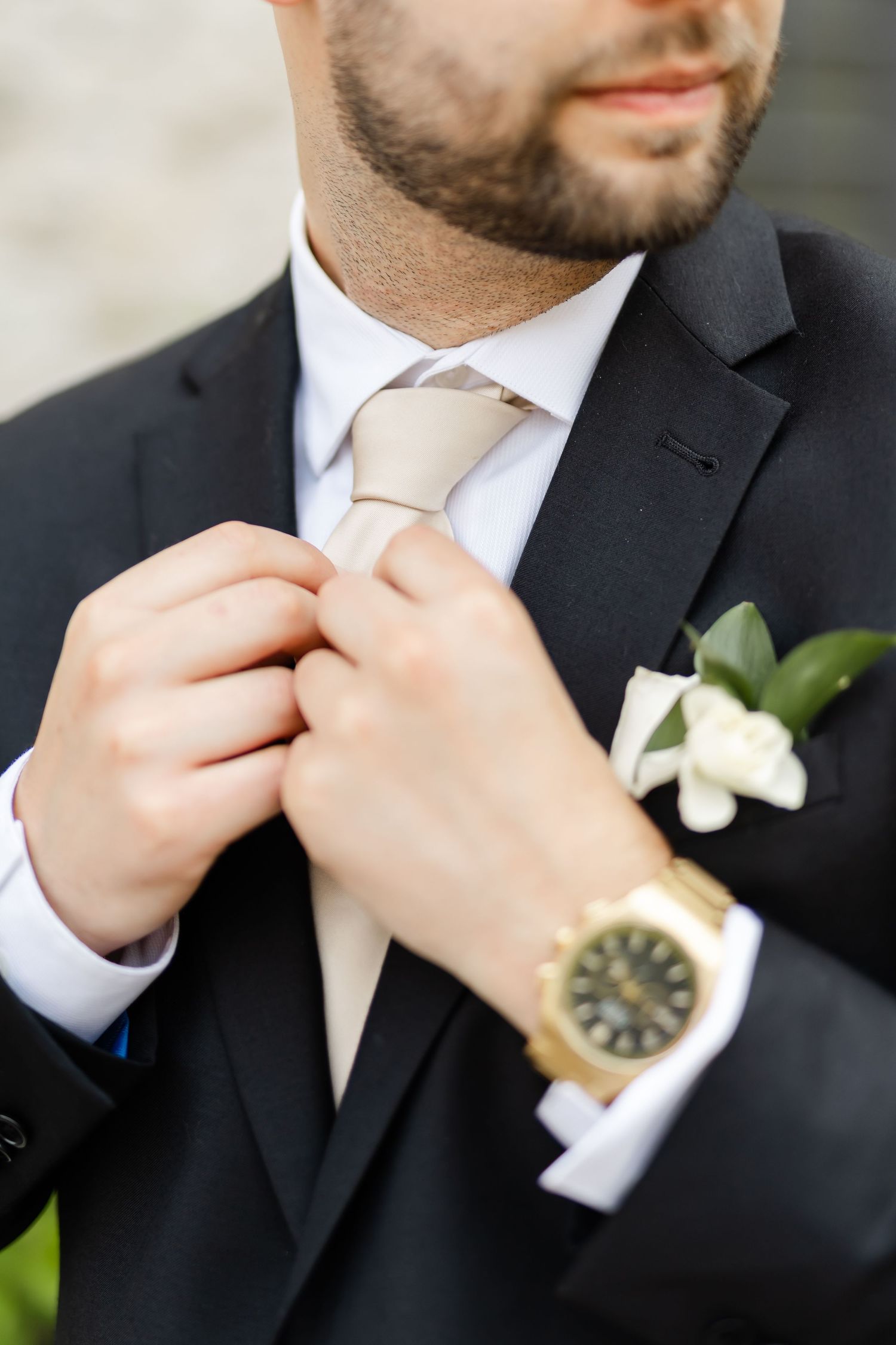 Groom in a black suit adjusts his champagne tie while wearing a gold Rolex watch and white boutonniere.