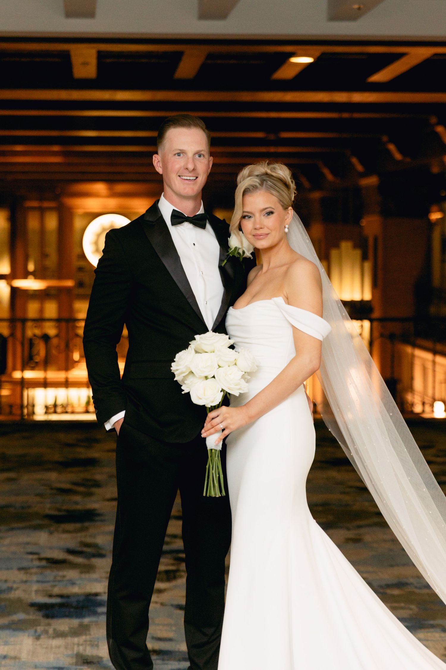 Bride and groom pose elegantly in a warmly lit venue, she in a white off-shoulder gown with veil, he in a black tuxedo.