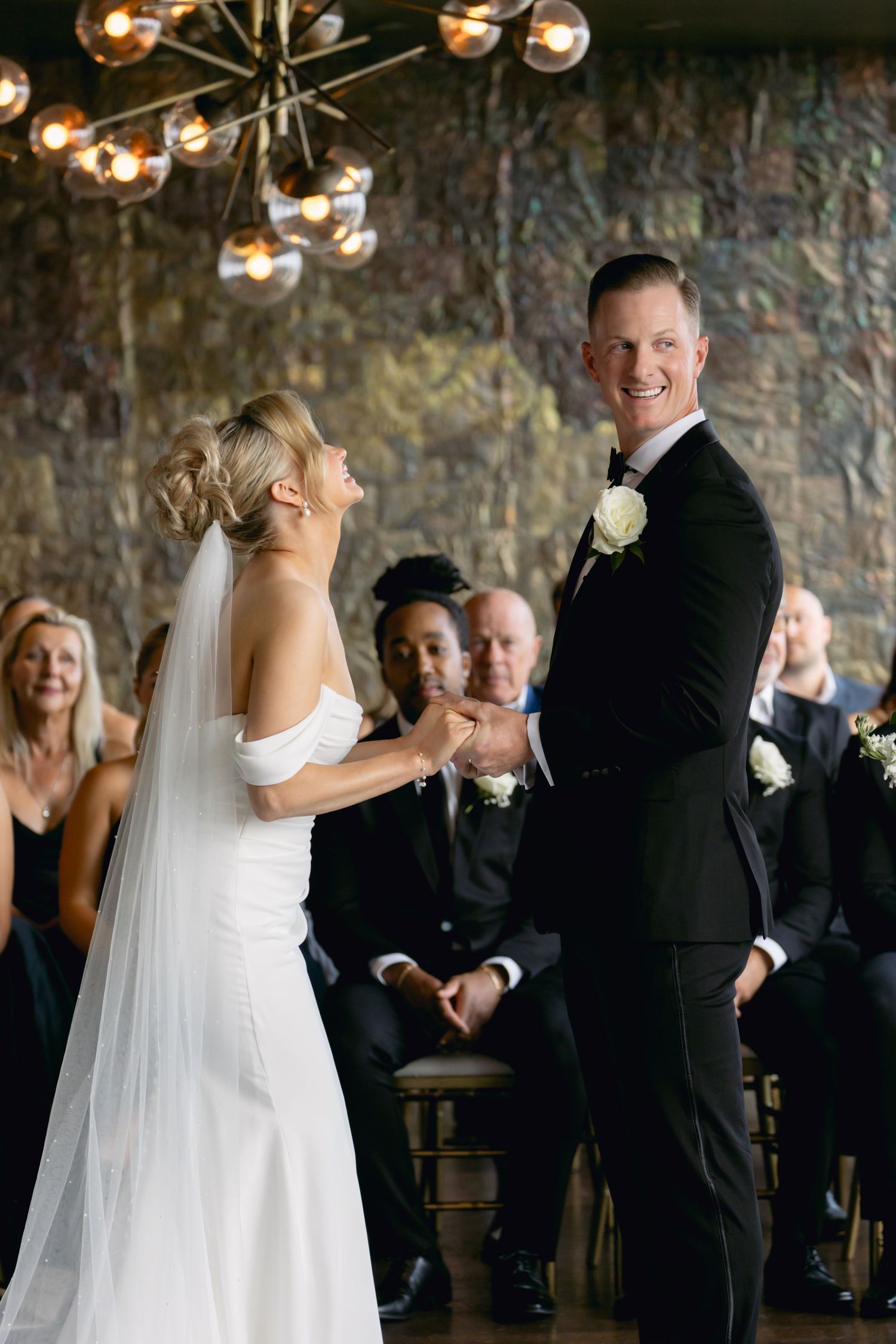 Smiling groom in black tuxedo holds bride's hands during indoor wedding ceremony with seated guests behind them.