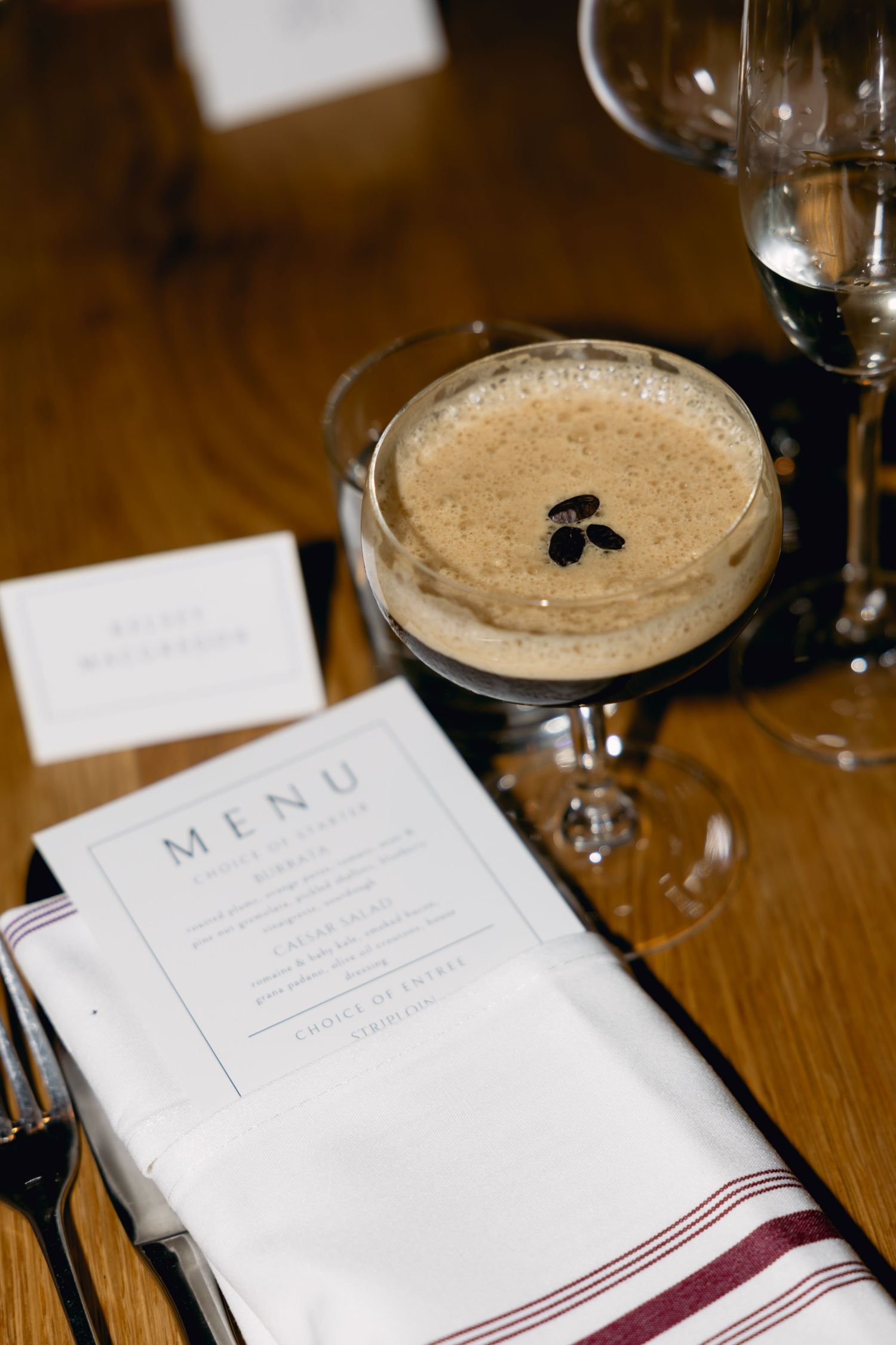 Elegant wedding table setting with espresso martini, menu card, and glassware on wood table.