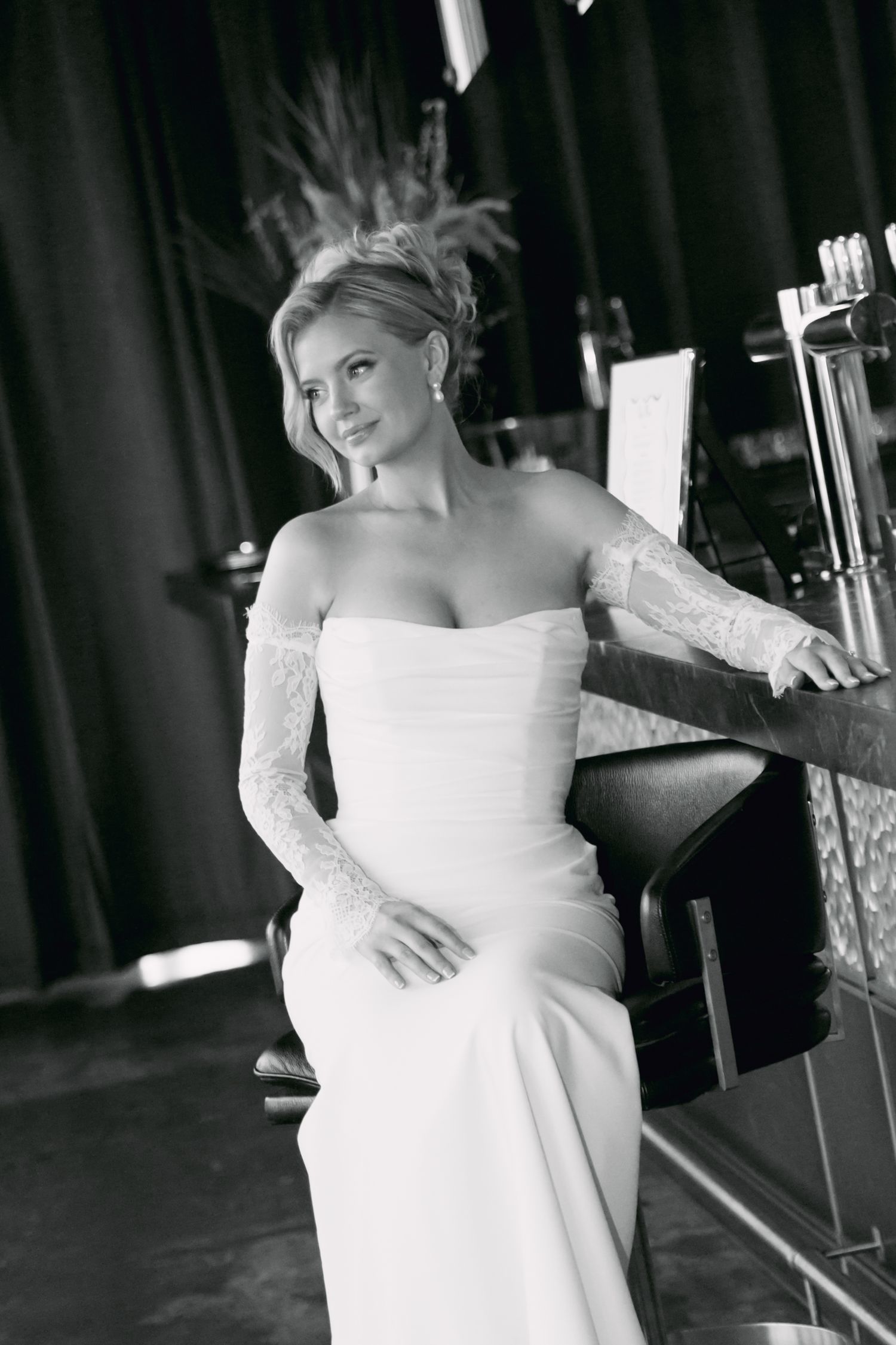 Black and white portrait of bride in white strapless gown with lace gloves seated at bar.
