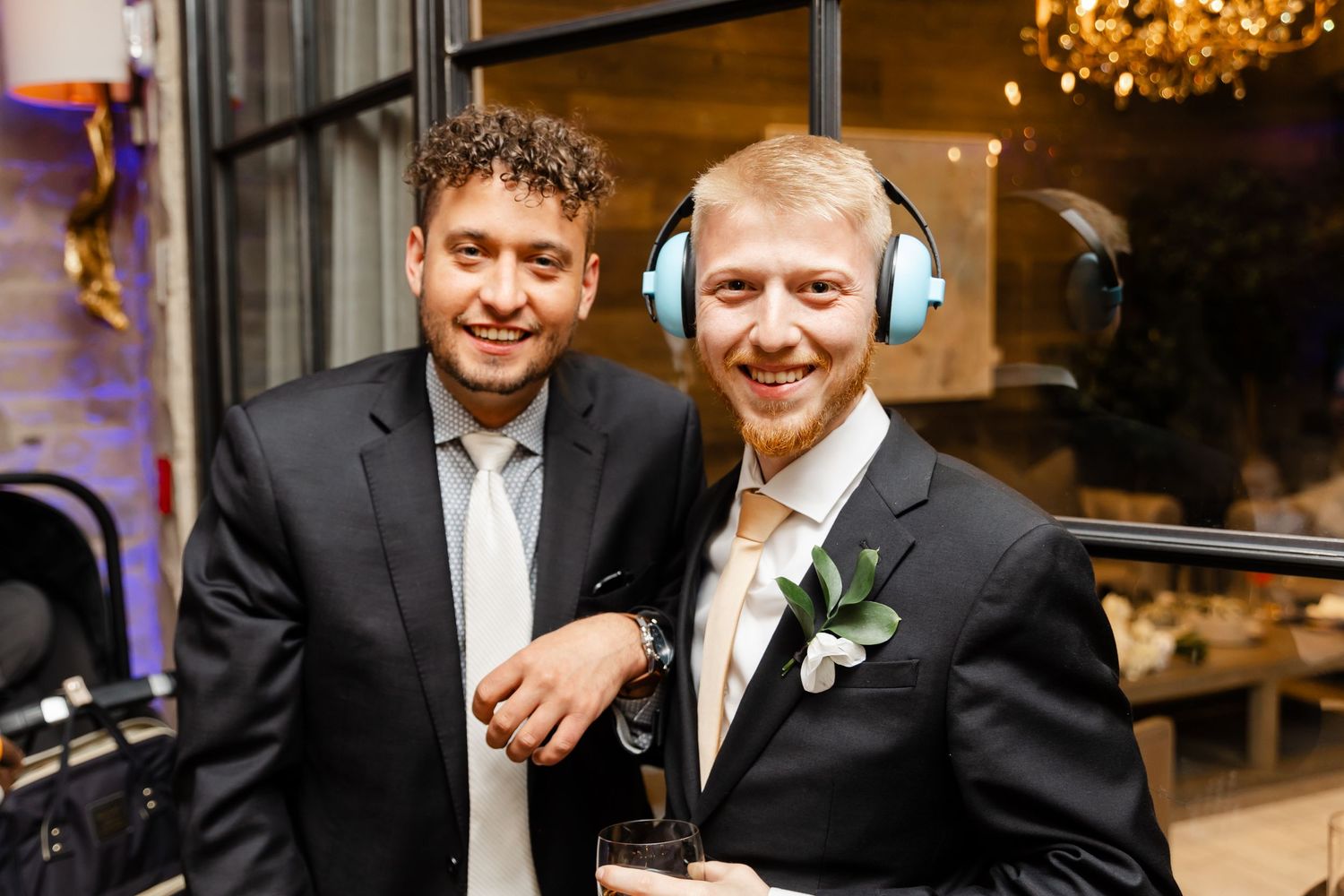 Two male wedding guests smile for photo, one wearing blue ear defenders at evening reception.