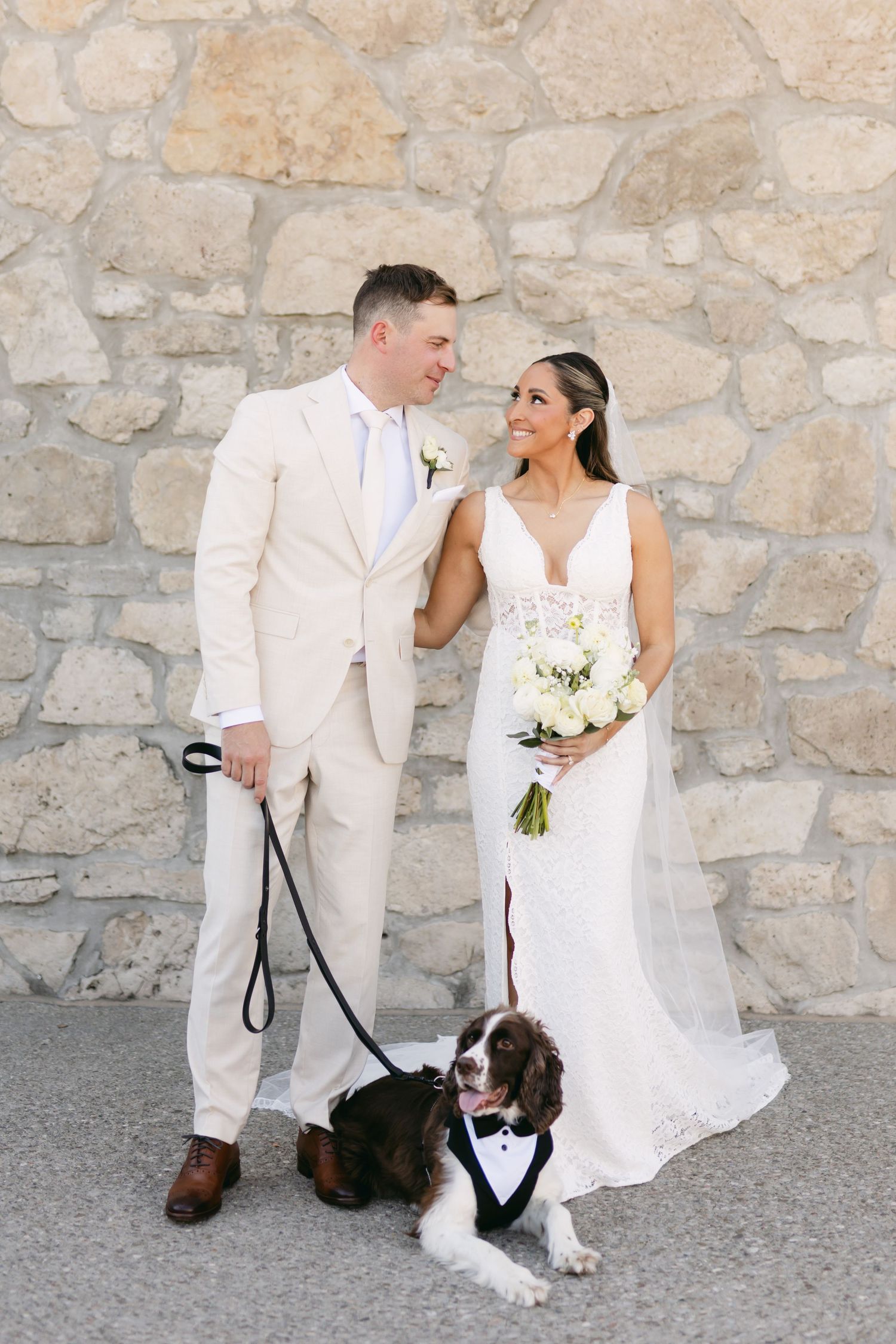 Wedding couple poses with their dog wearing a tuxedo bow tie against a stone wall.