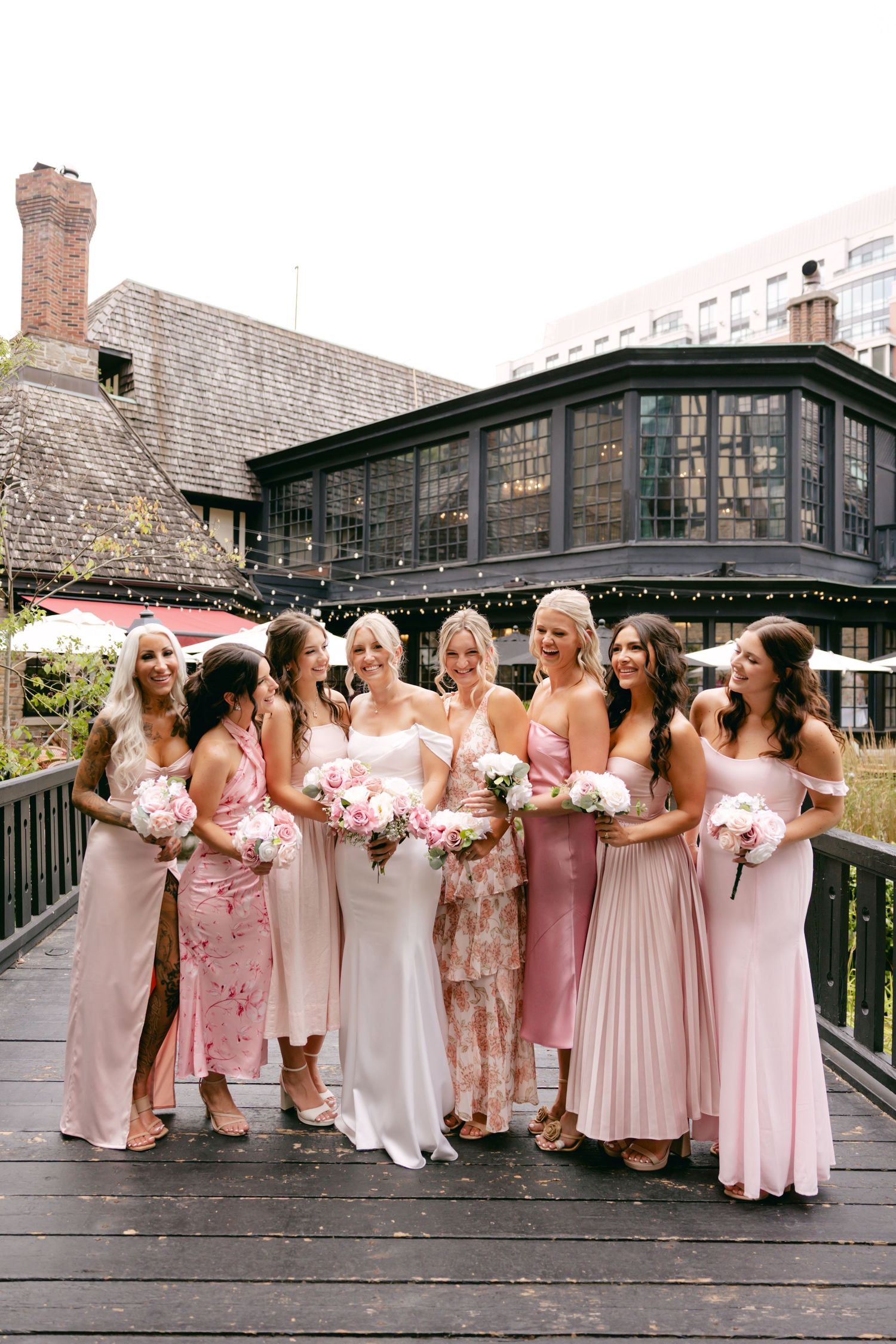 Bridesmaids in pink dresses pose together on wooden deck during wedding celebration.