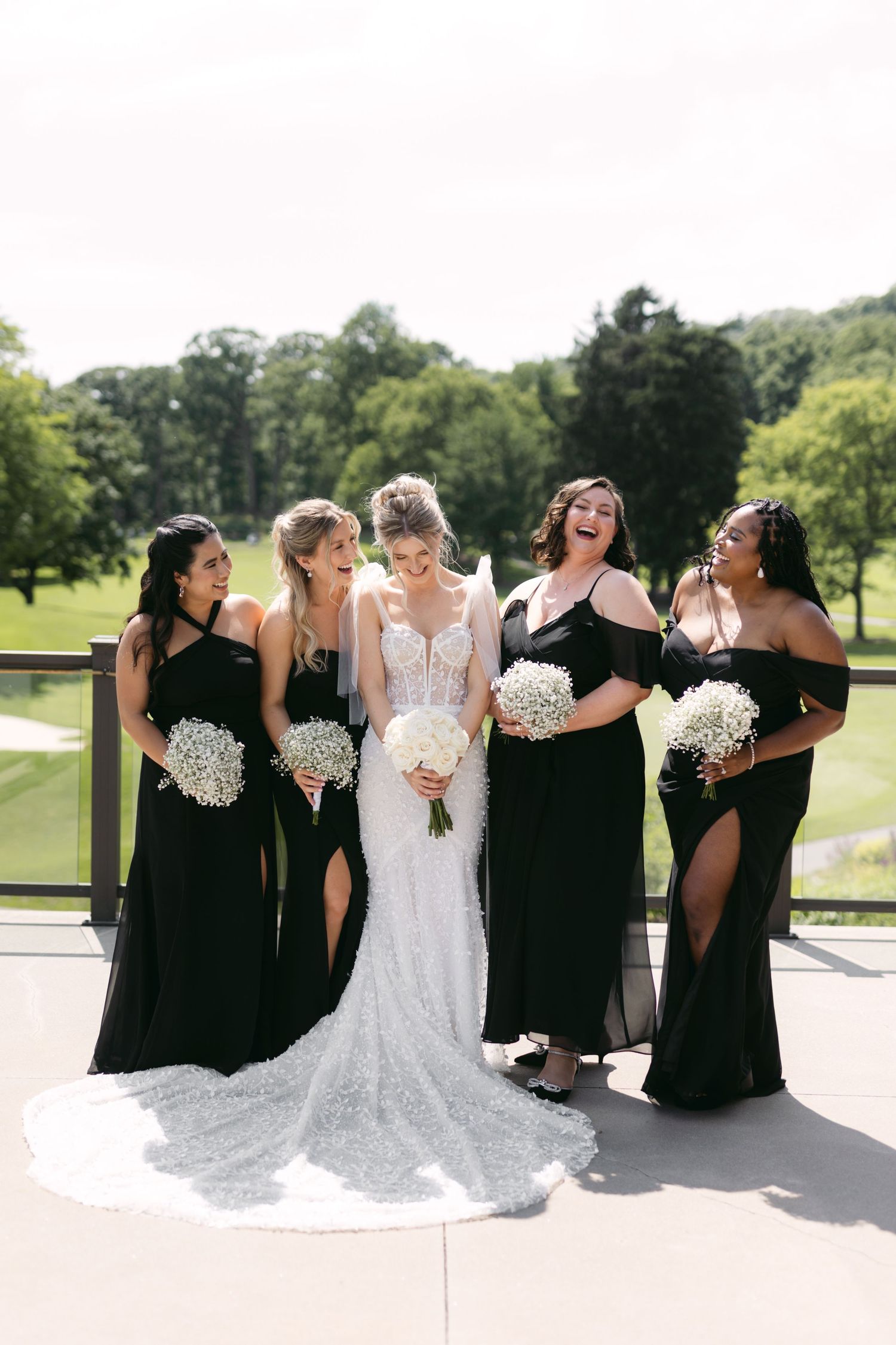 Bridal party laughs together while holding white bouquets in black dresses and white wedding gown.
