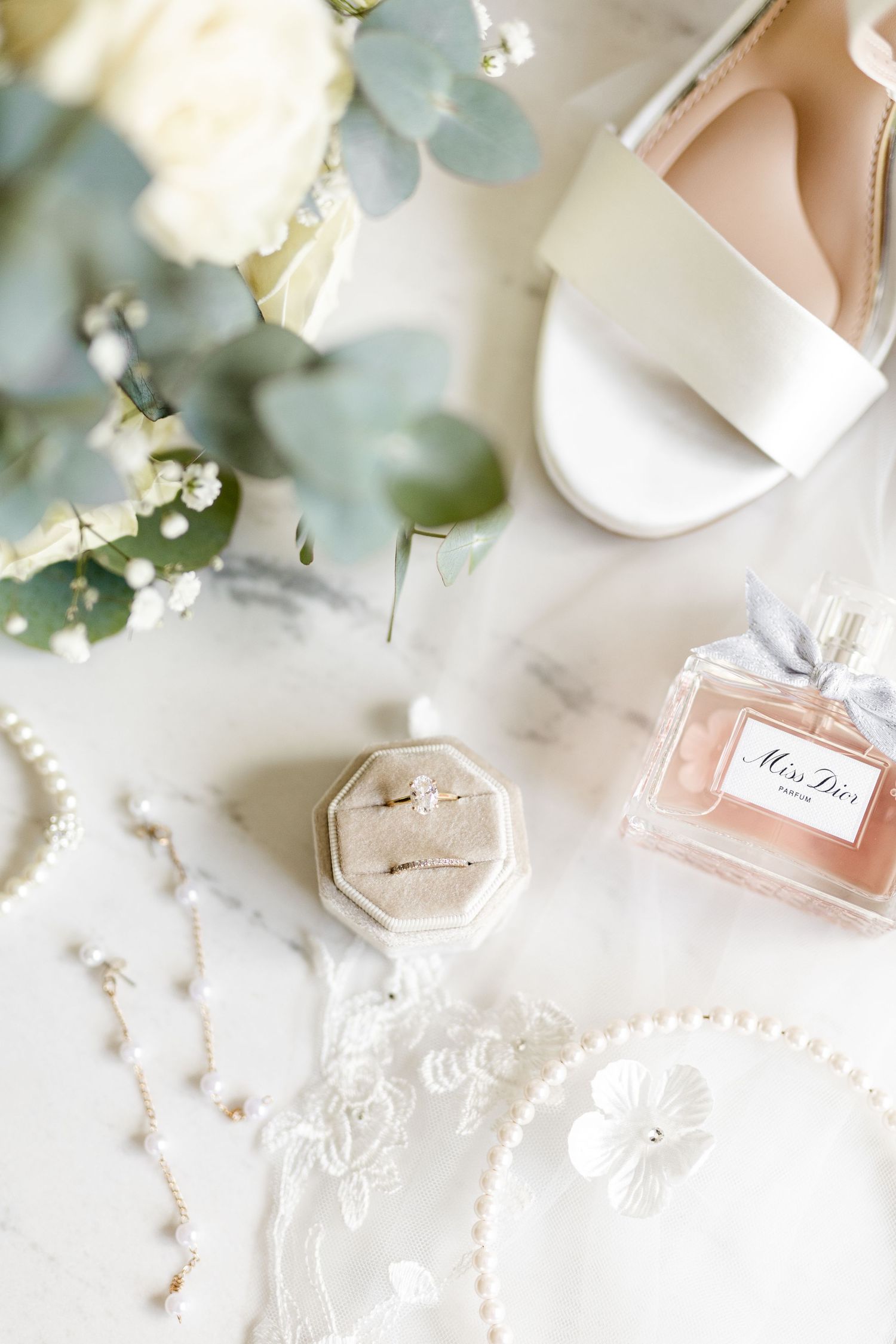 Wedding details flatlay with perfume, rings, pearls and blush shoes on marble surface.