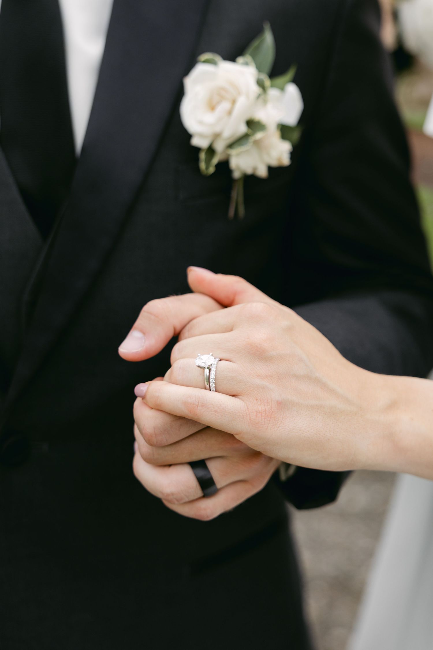 Close-up of clasped hands showing wedding rings and a white rose boutonniere on a black tuxedo lapel.