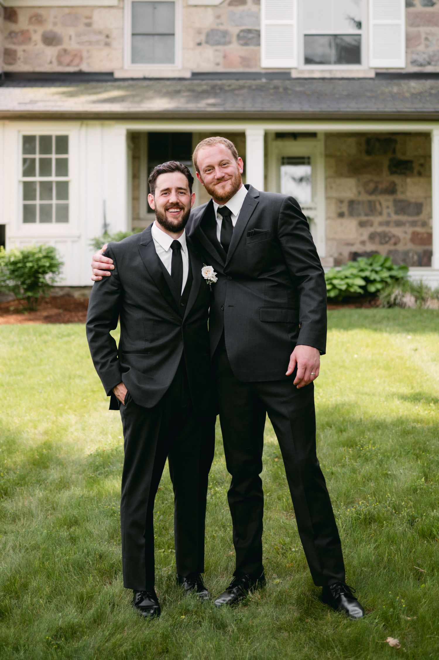 Groomsmen in black tuxedos pose playfully on a grassy lawn outside a stone building.