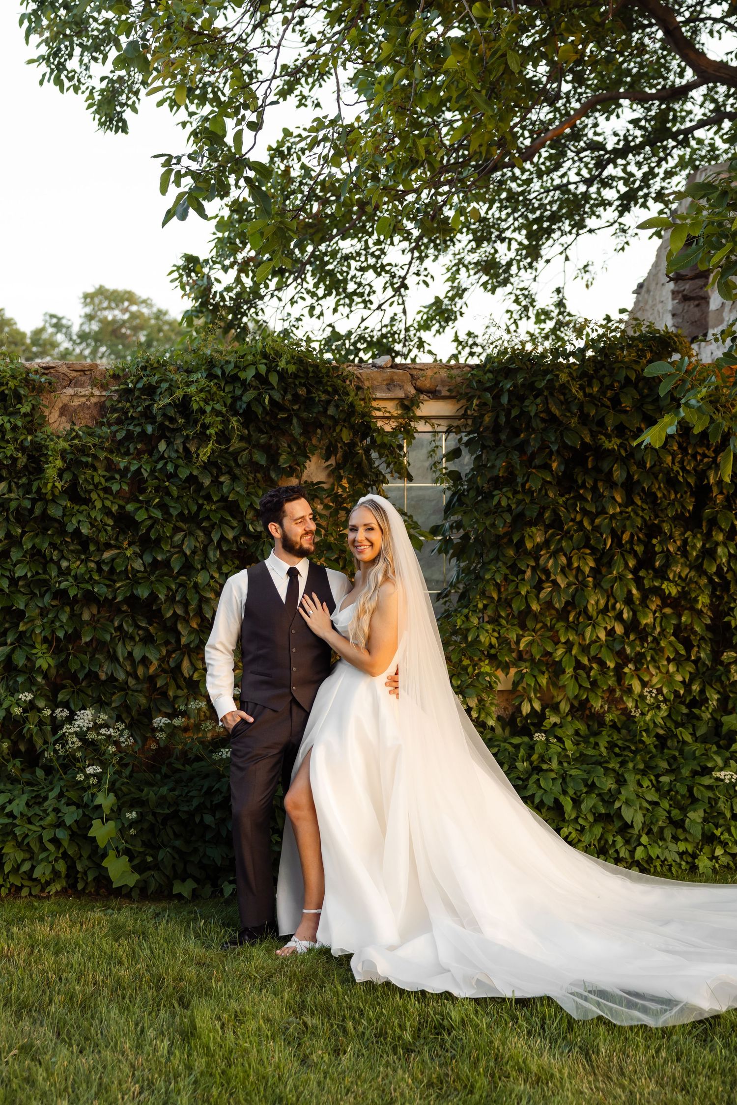 Bride in flowing white gown poses with groom against an ivy-covered wall at sunset.