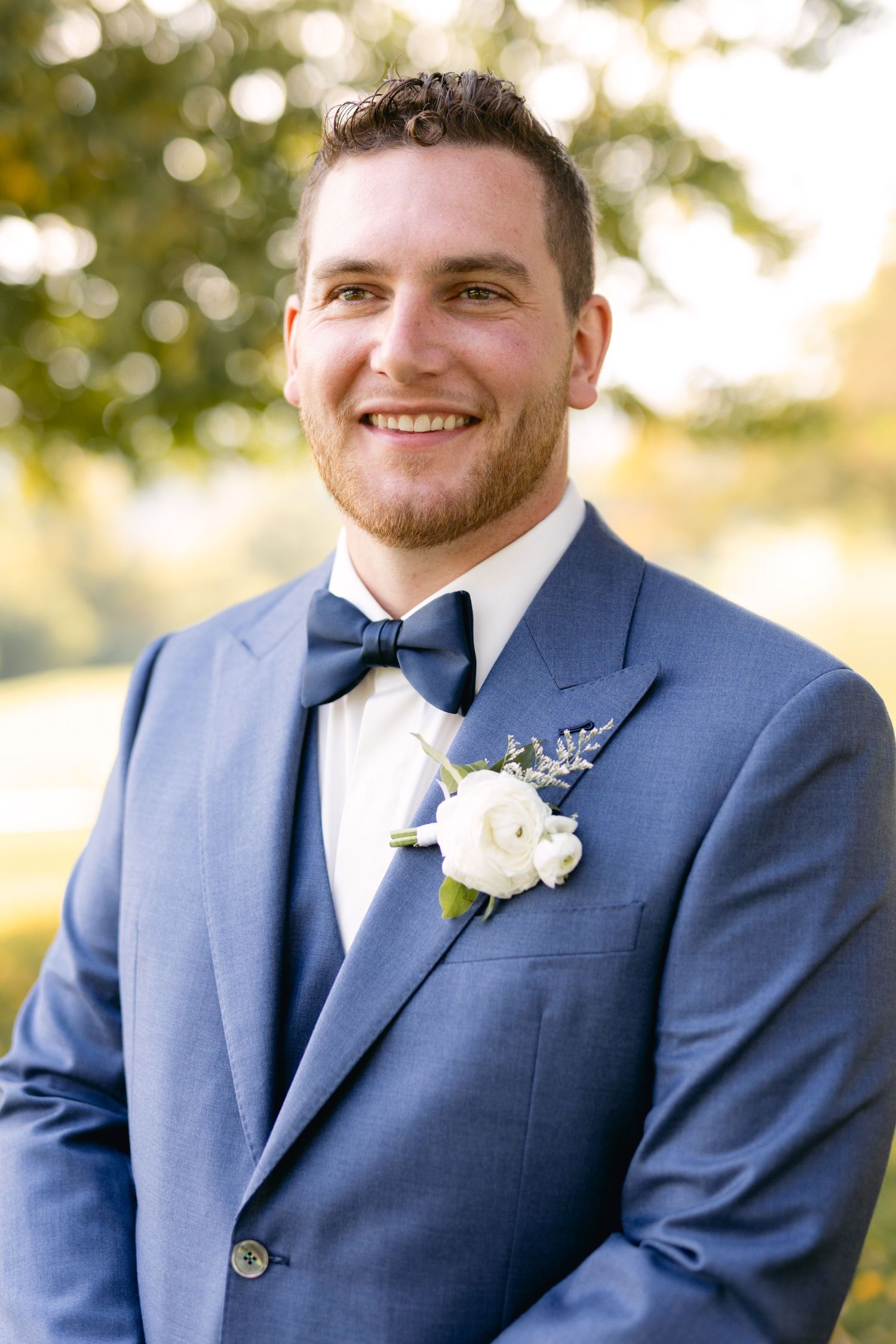 Groom wearing navy suit and bow tie smiles at camera during outdoor wedding portraits.