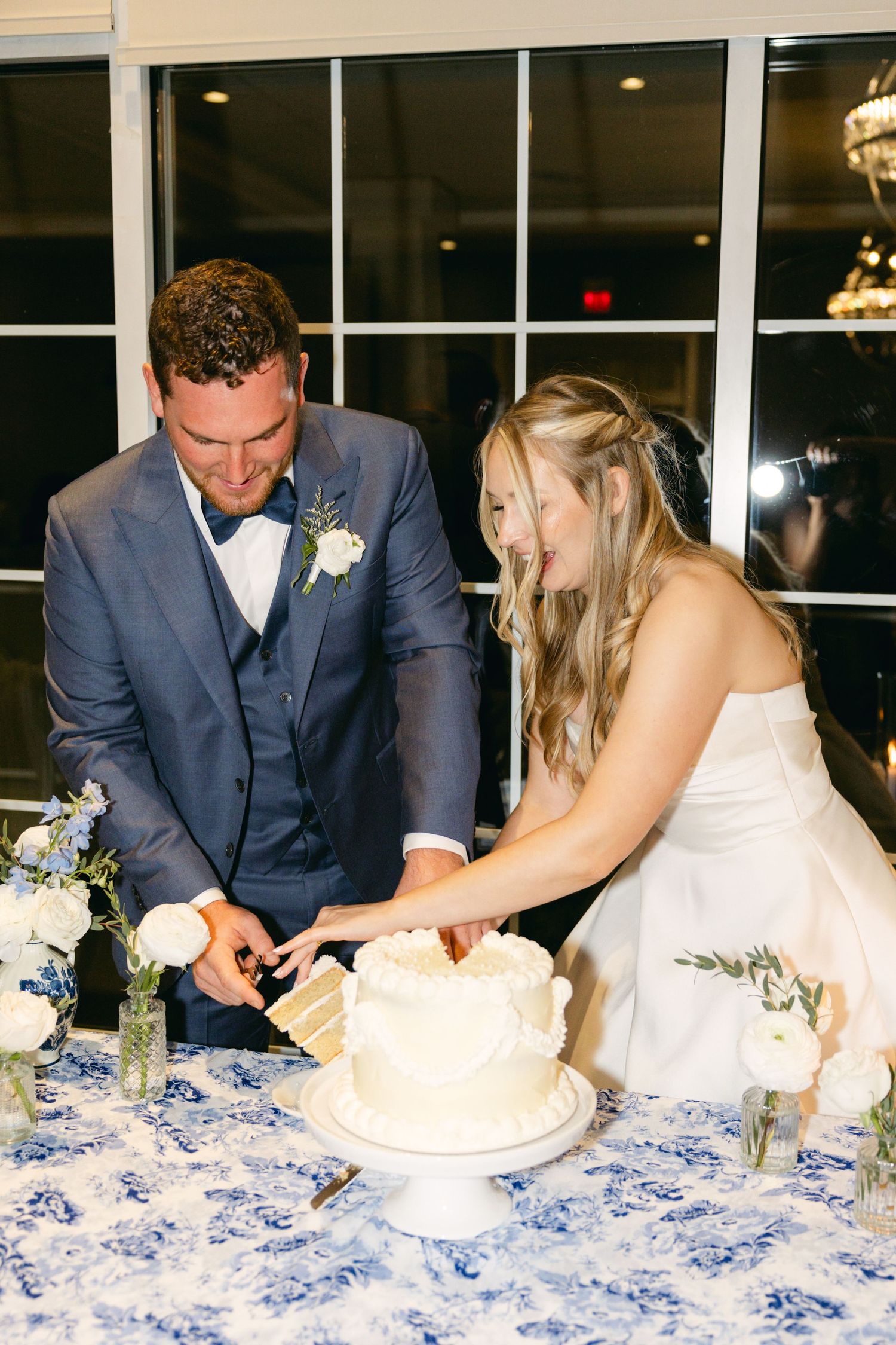Newlyweds cut their white wedding cake together on a blue and white patterned tablecloth at their reception.