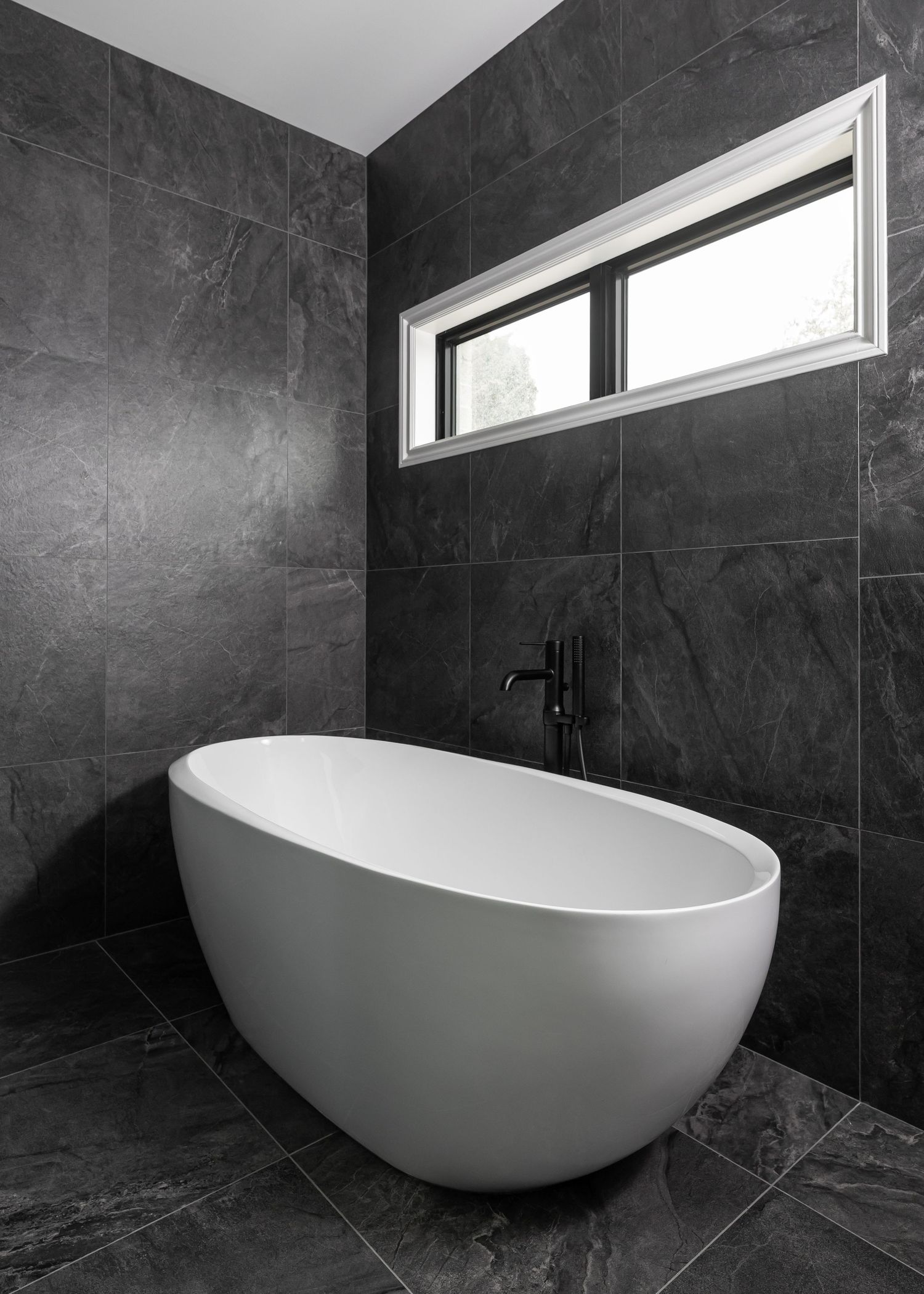 Contemporary white freestanding egg-shaped bathtub against dark marble tile walls.