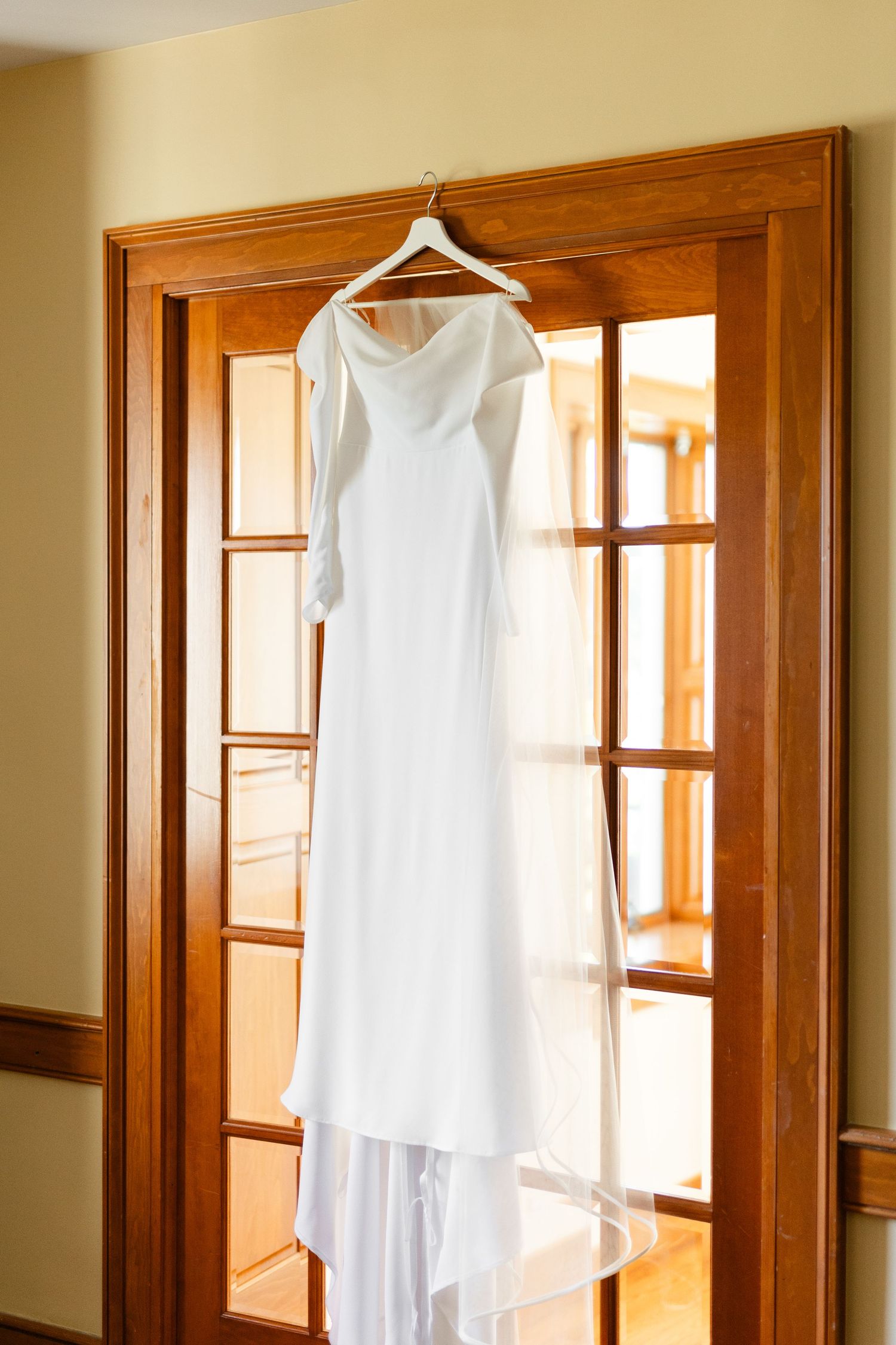 An elegant white wedding dress hangs on display against wooden French doors.