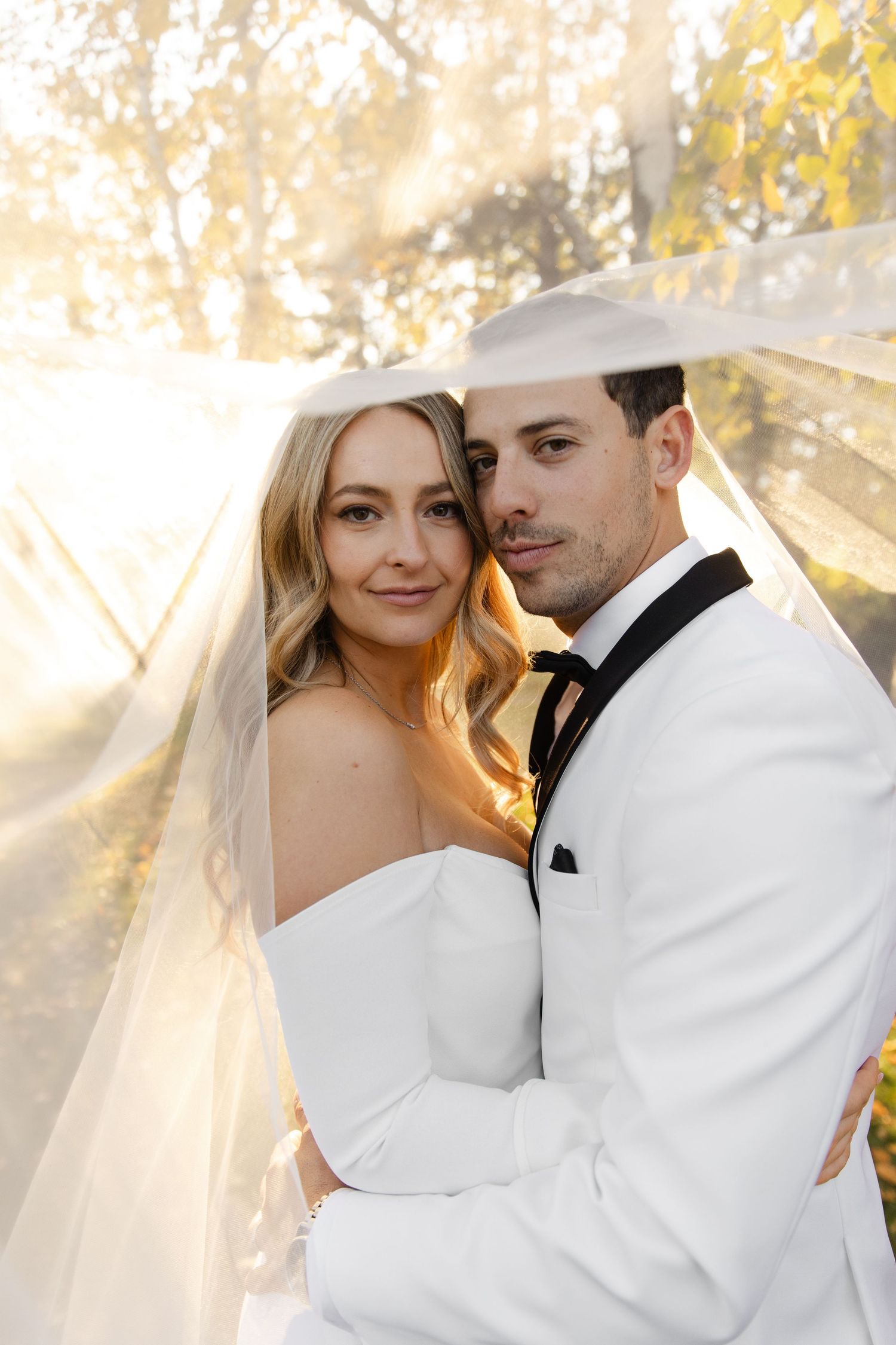 Newlyweds share intimate moment in autumn setting with bride in white gown and groom in white tuxedo.