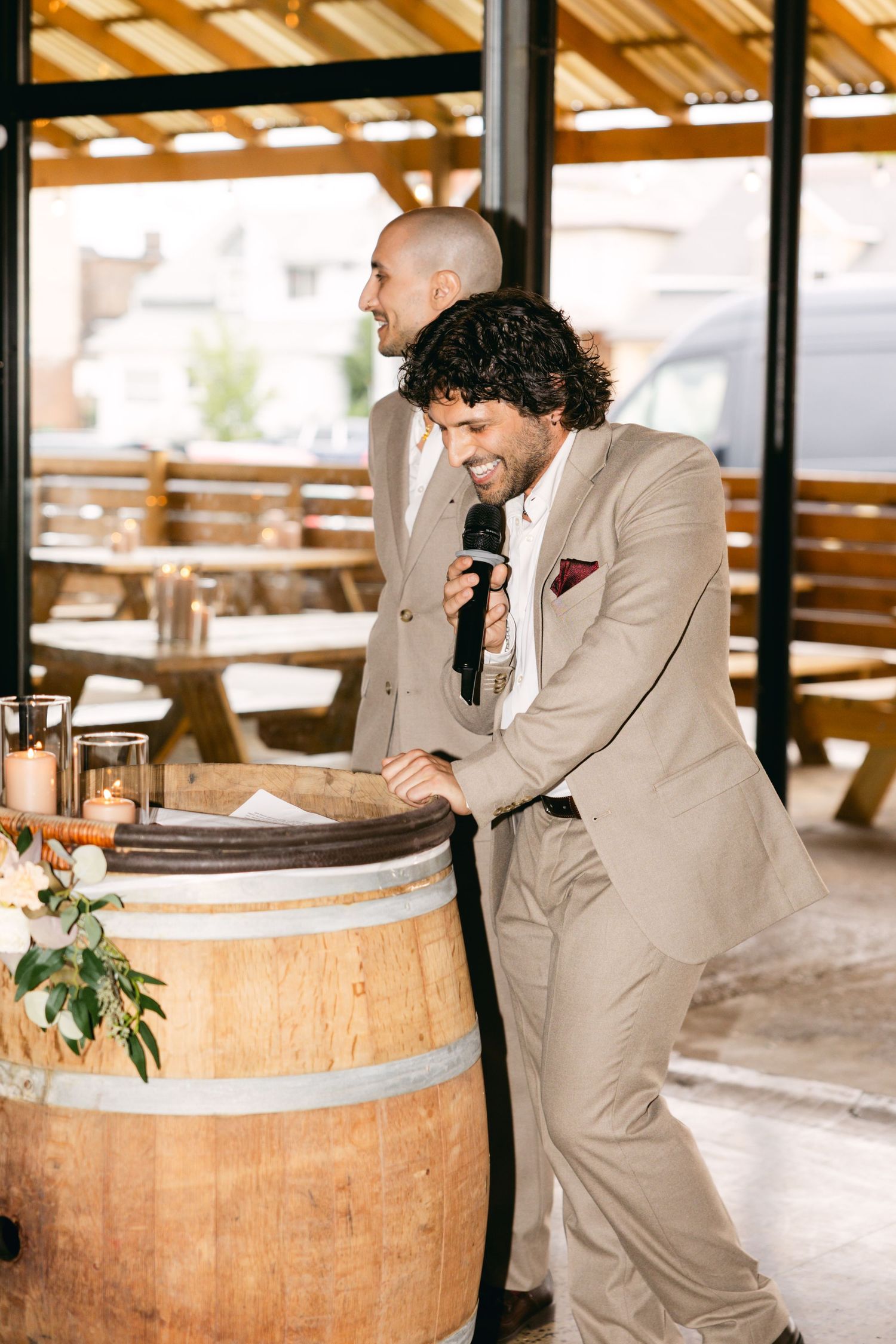 Groomsmen share laughter while leaning on wooden barrel at rustic wedding venue.