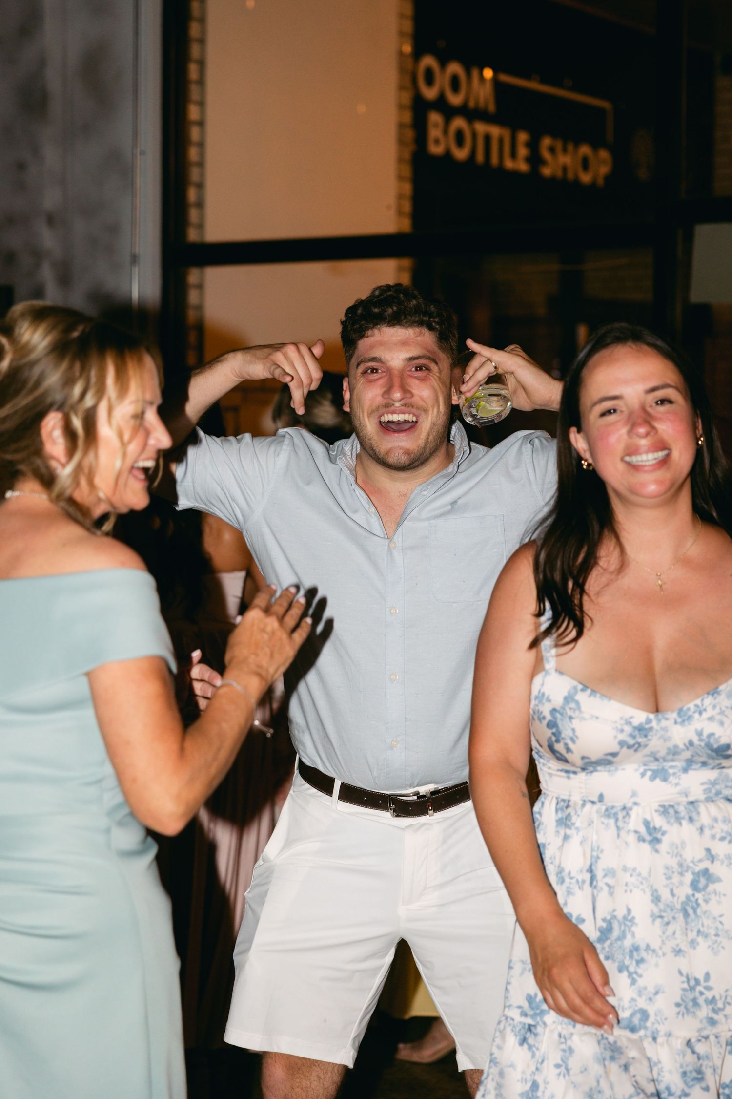 Wedding party dancing and laughing together at a reception with a bottle shop sign visible.