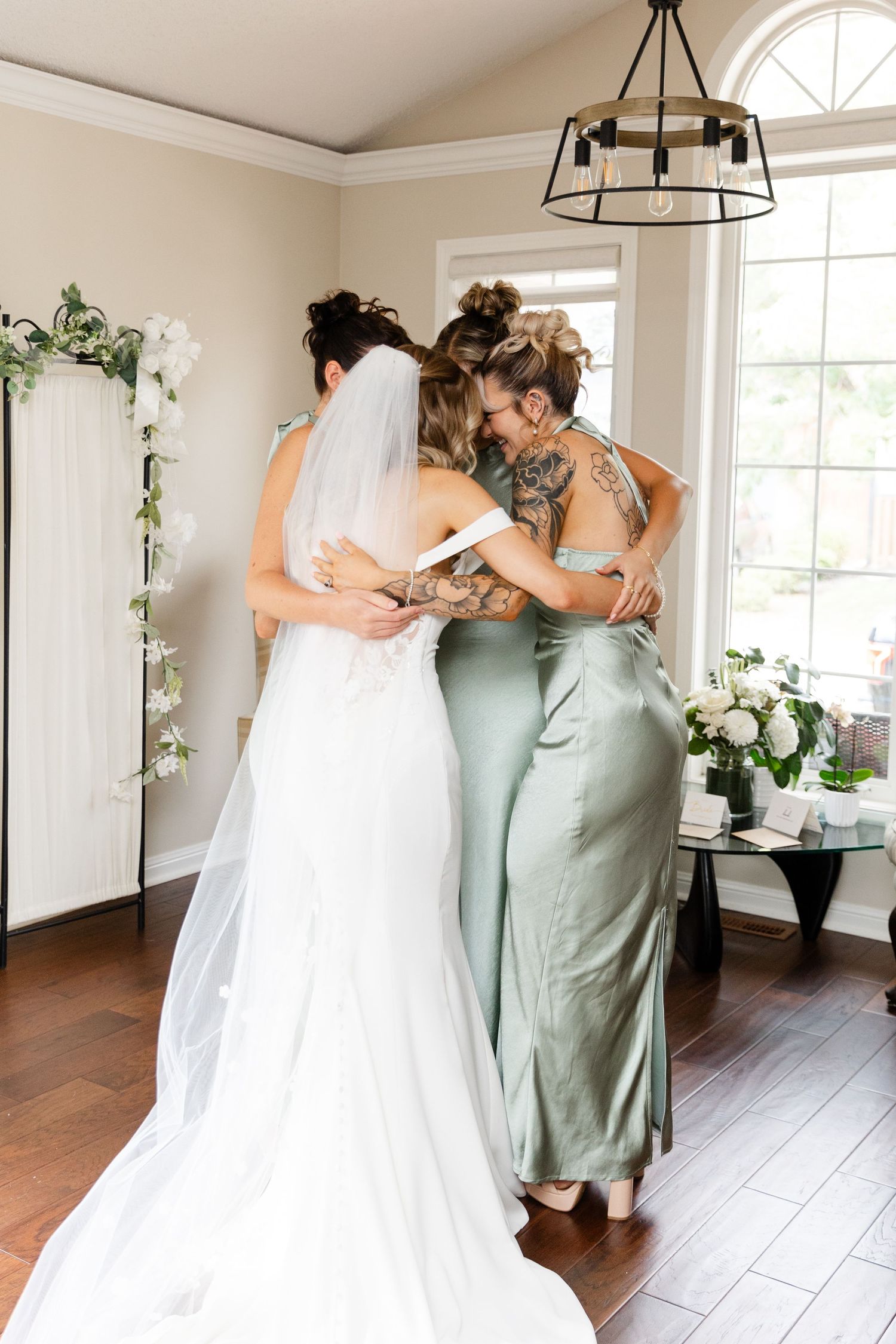 Wedding party members in sage green dresses gather around the bride in her white gown.
