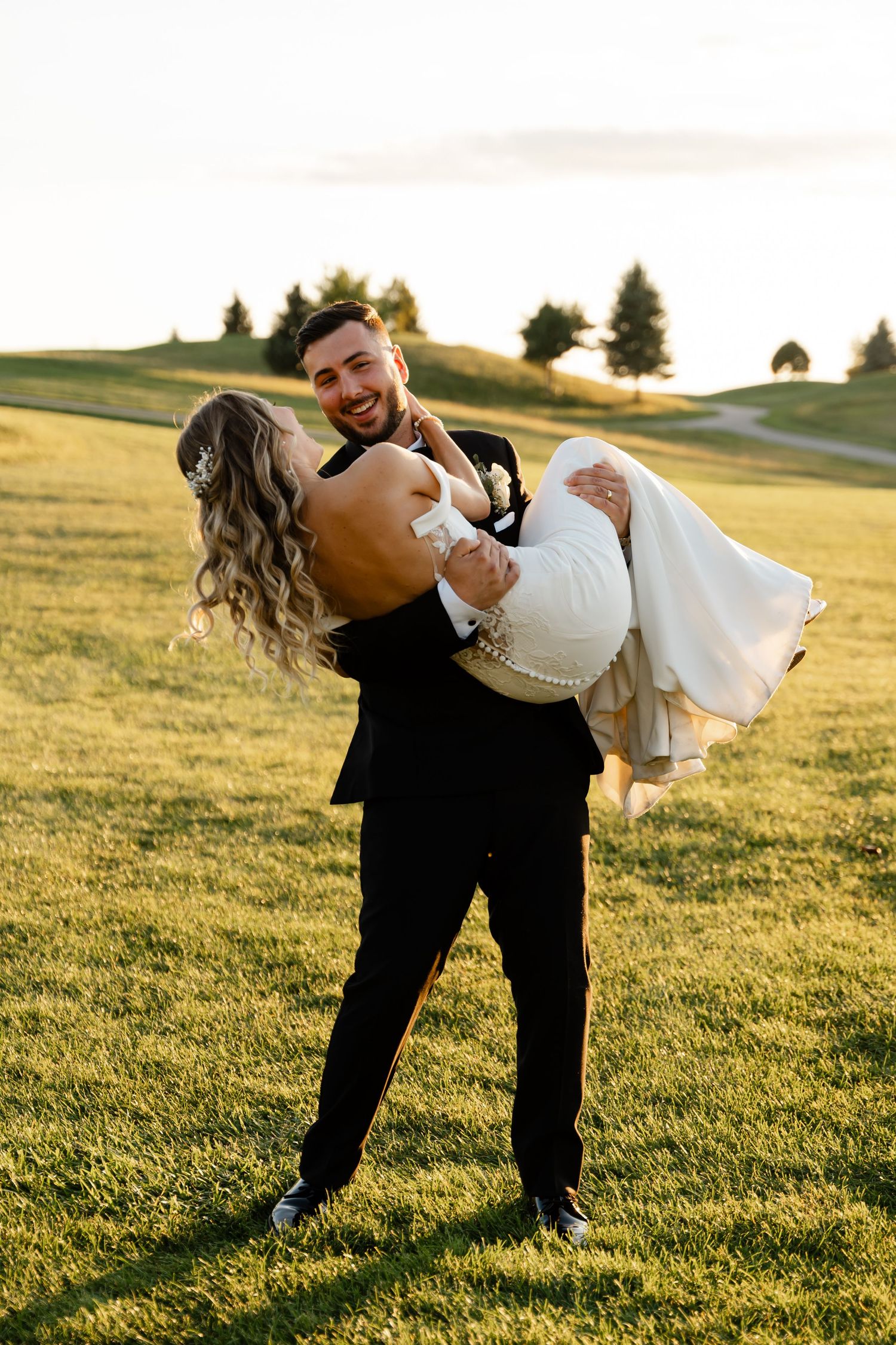 A romantic wedding photo of a couple on a grassy hill at sunset with the groom carrying the bride.