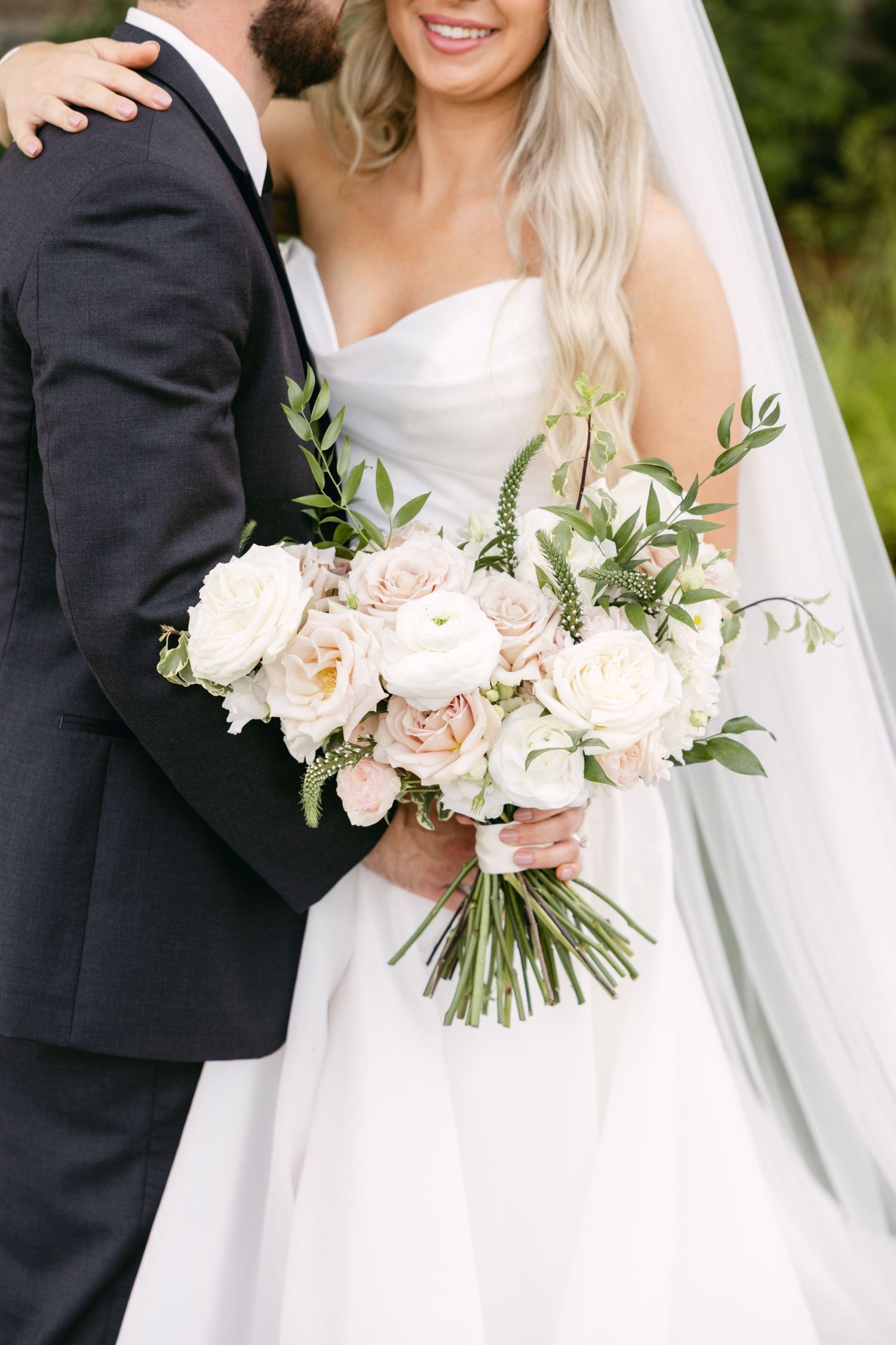 Wedding couple embraces while holding elegant white and blush floral bouquet.