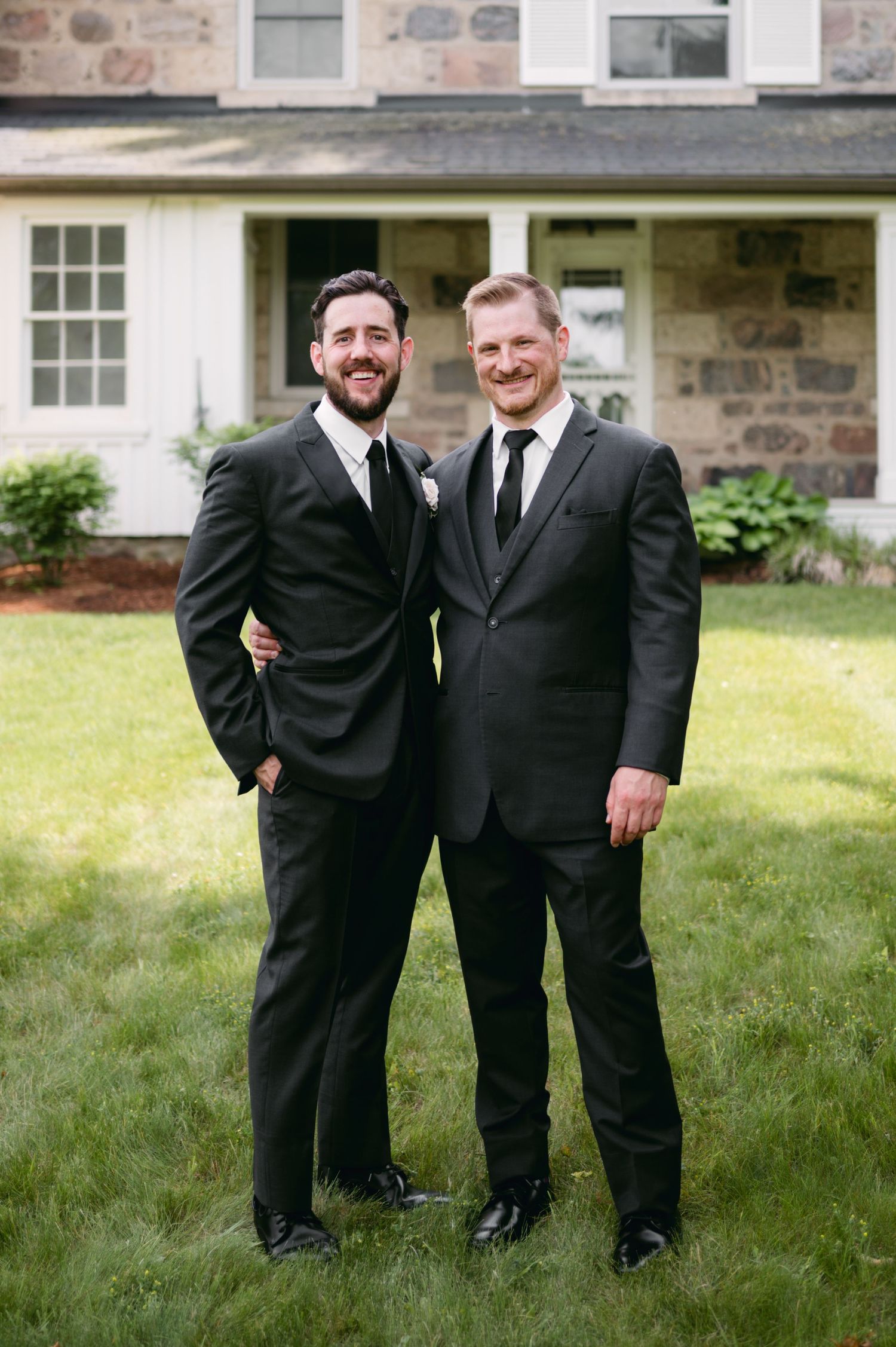 Two groomsmen in black suits share candid moment on lawn by stone building.