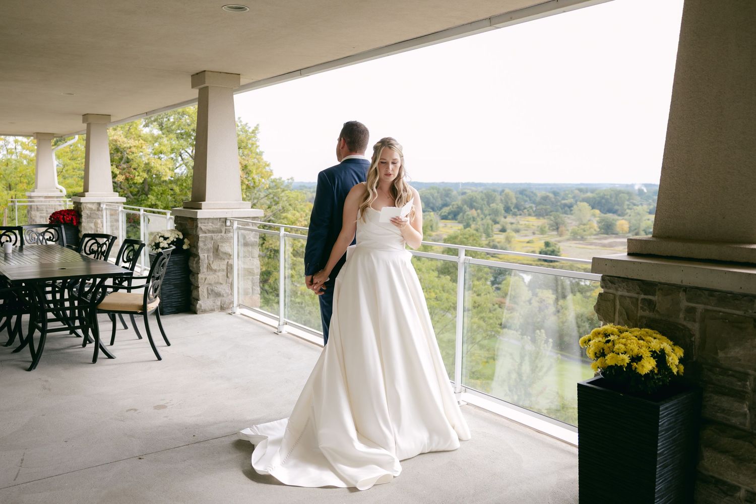 A couple in wedding attire stands together on an elegant covered balcony with scenic views.