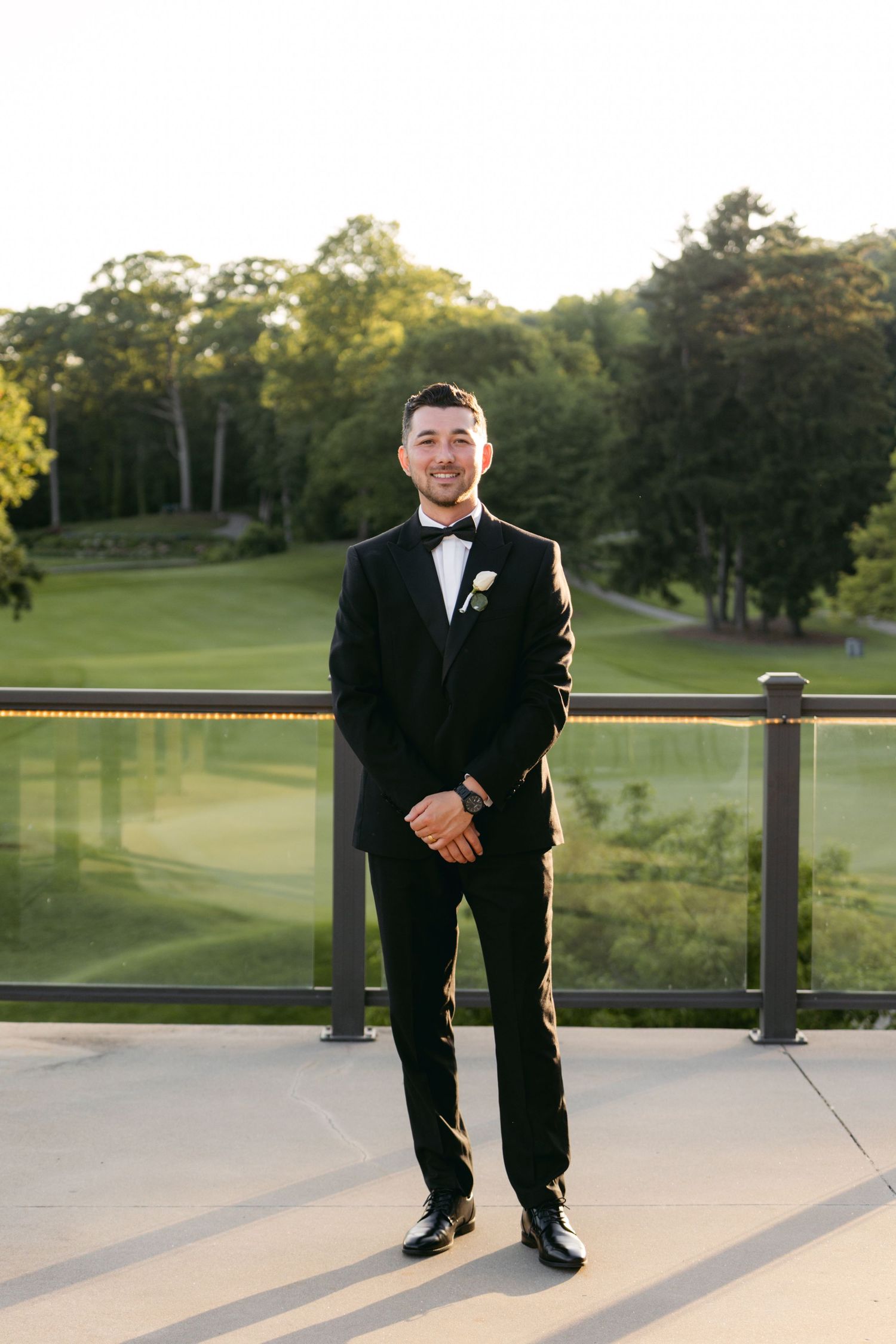 Groom in classic black tuxedo poses on outdoor terrace with golf course scenery.
