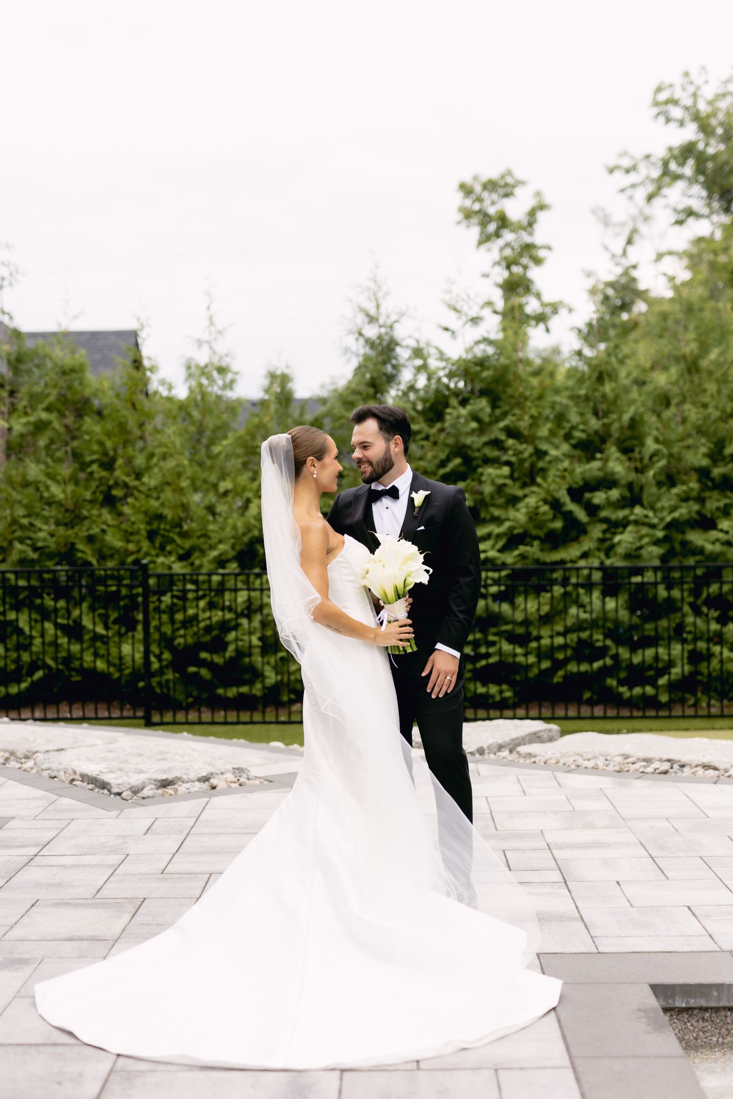 Bride and groom pose outdoors in garden setting, bride in flowing white gown with long veil.