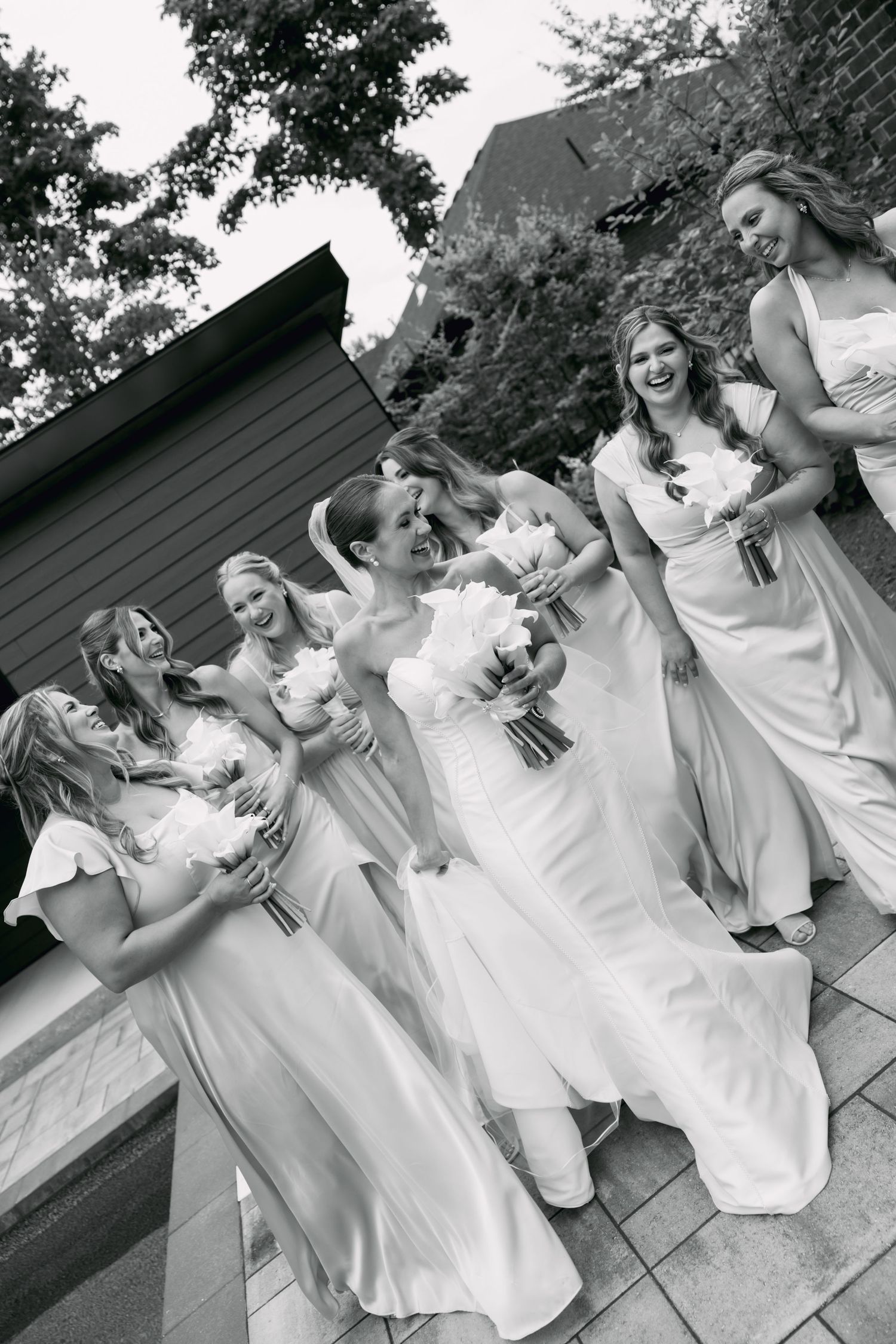 Black and white photo of bride with bridesmaids in long gowns laughing outdoors at wedding.