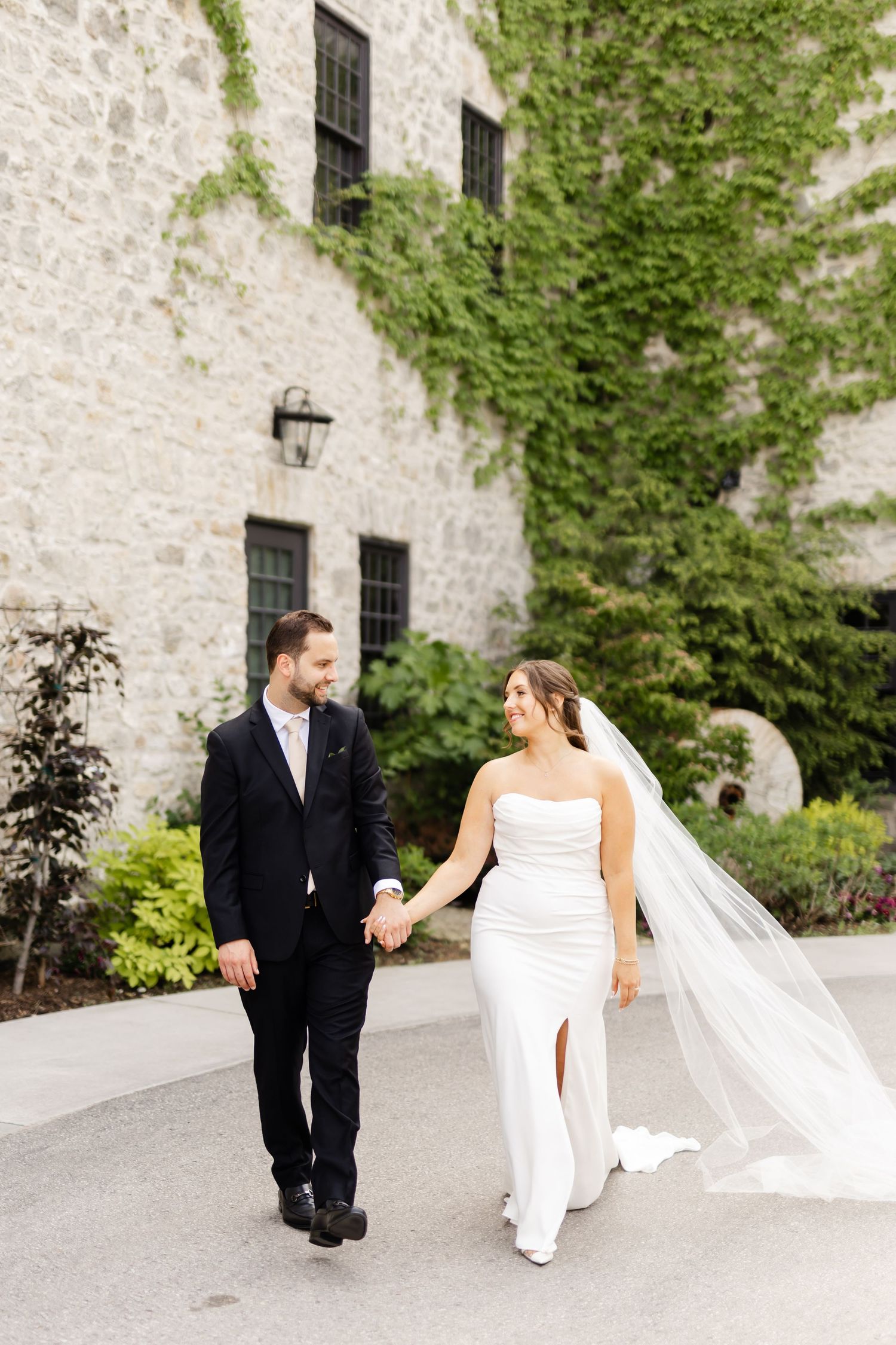 Bride and groom walk hand-in-hand past ivy-covered stone building, bride in white gown with flowing veil.