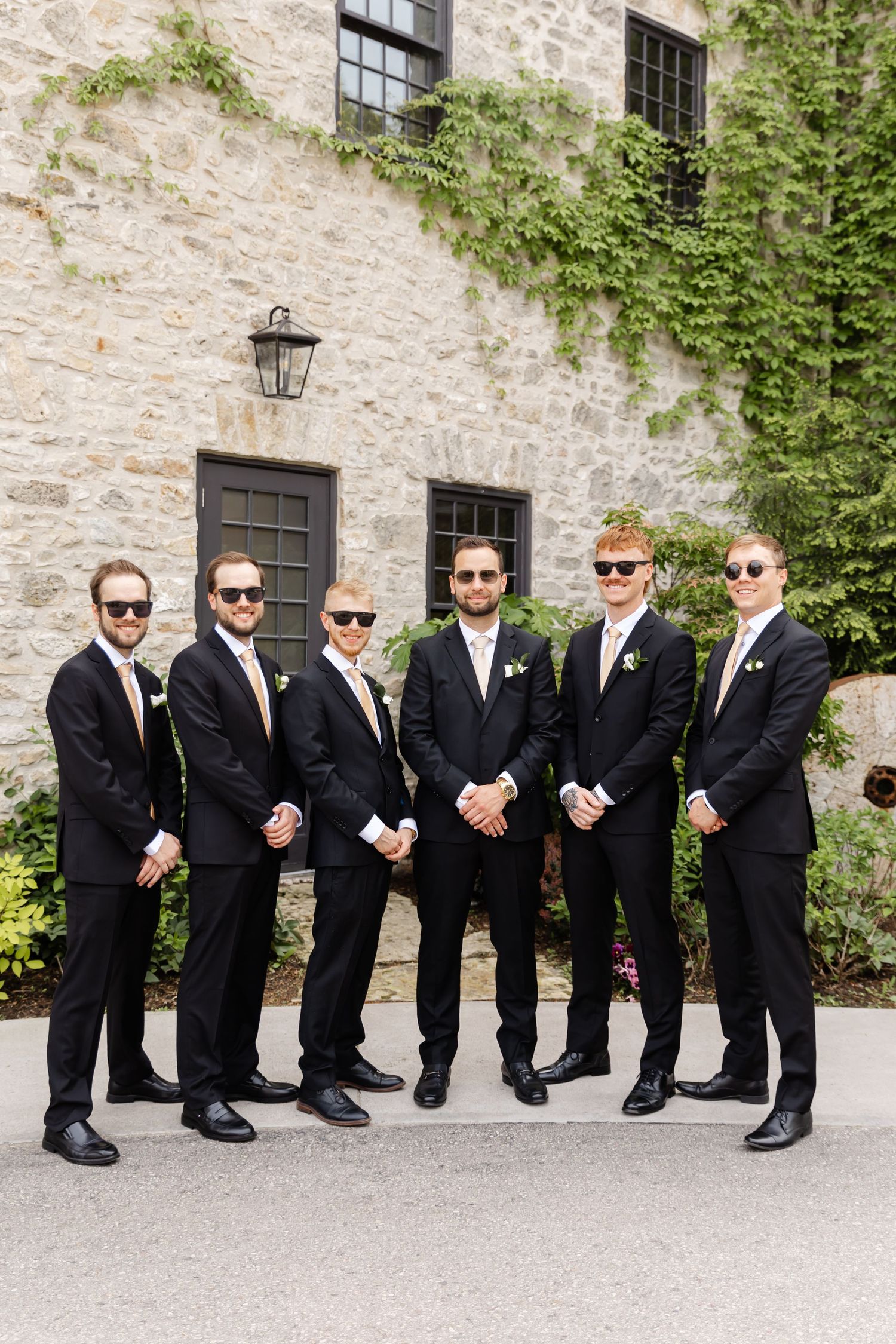 Six groomsmen in matching black suits and sunglasses pose outside stone building covered in ivy.