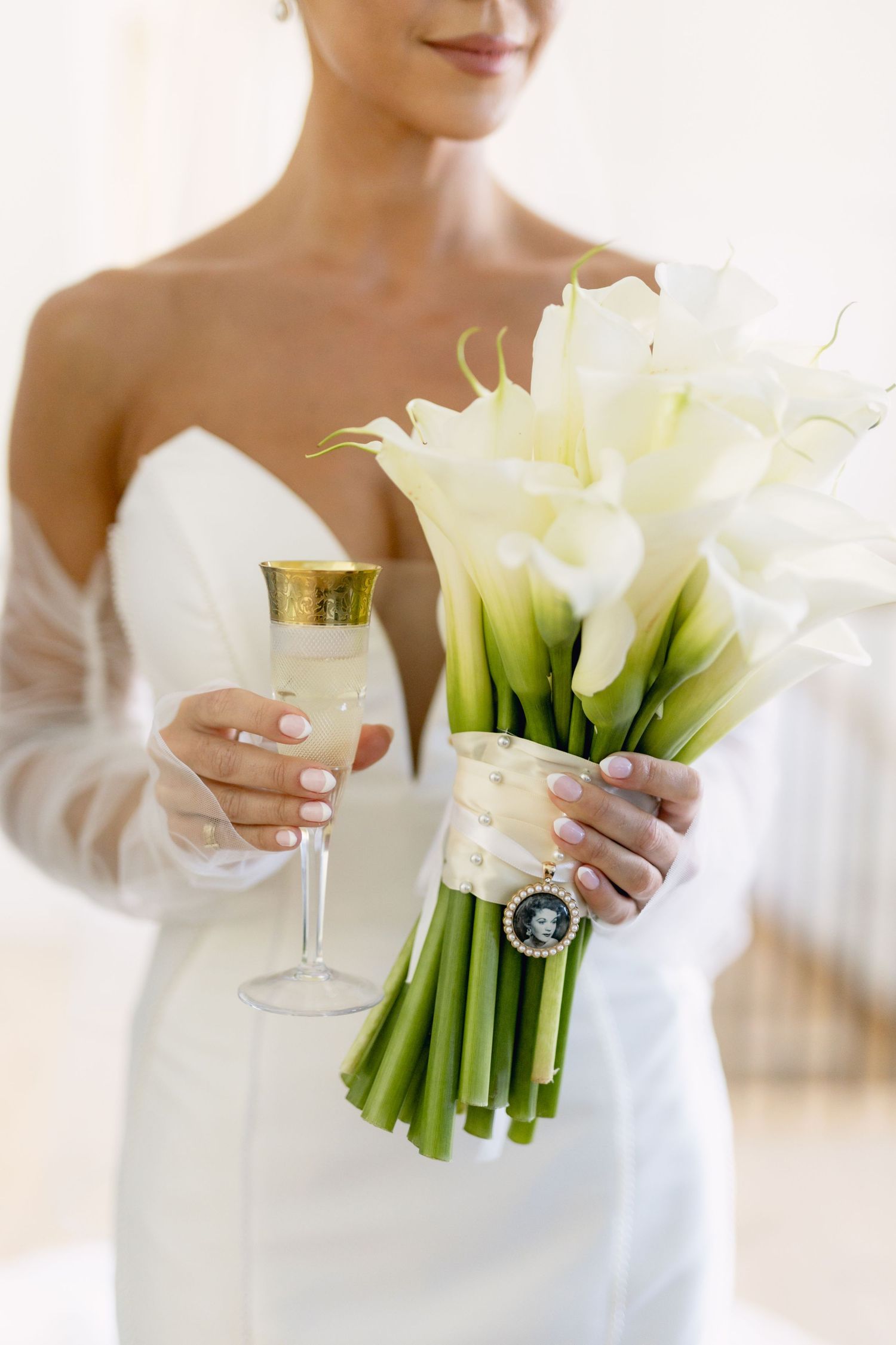 Bride in white gown holding calla lily bouquet and champagne glass with gold rim on her wedding day.