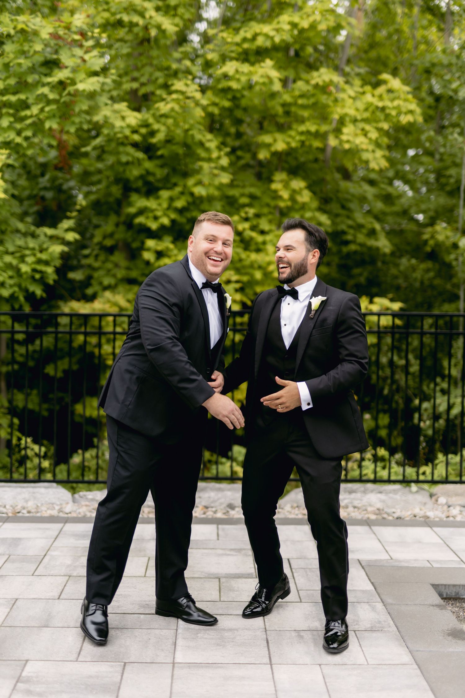Two groomsmen in black tuxedos laughing and shaking hands outdoors at a wedding.