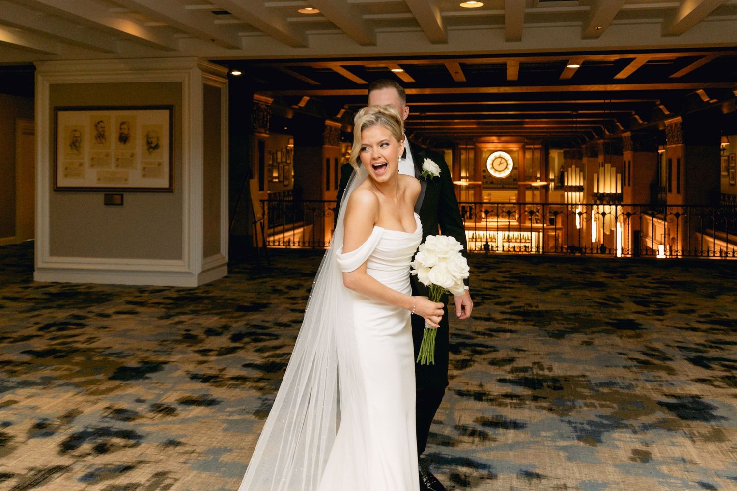 Bride in white off-shoulder gown holding bouquet with groom in tuxedo at elegant wedding venue.
