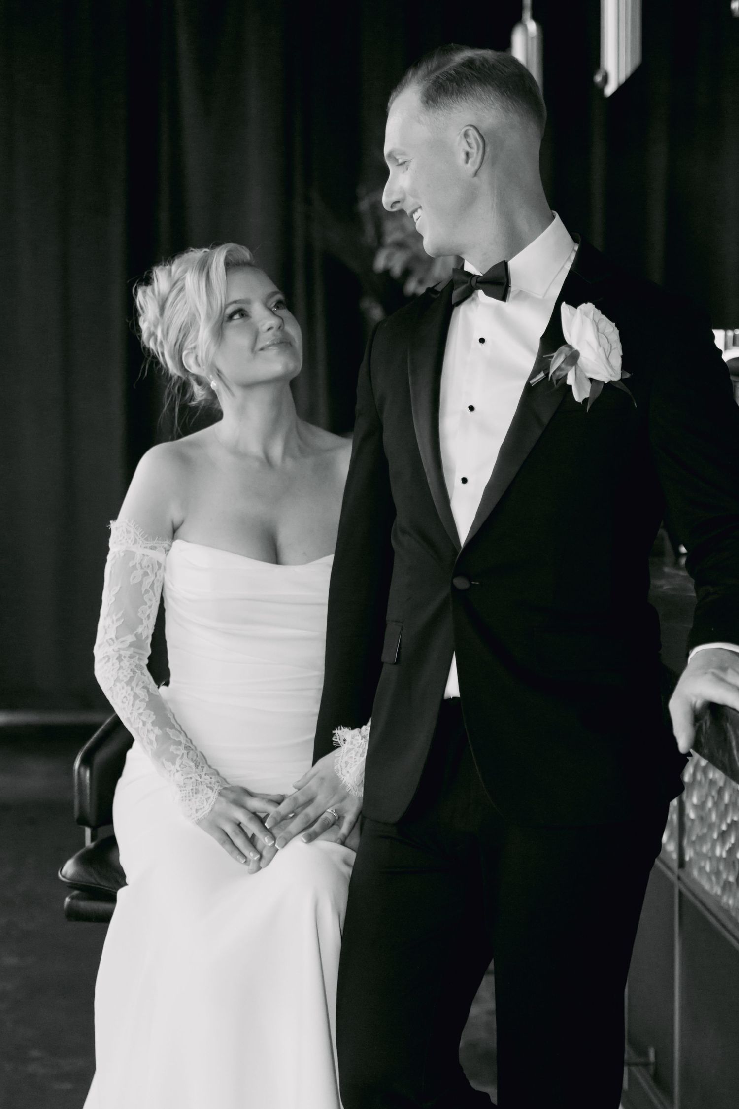 Black and white portrait of bride in off-shoulder gown gazing up at groom in bow tie tuxedo.