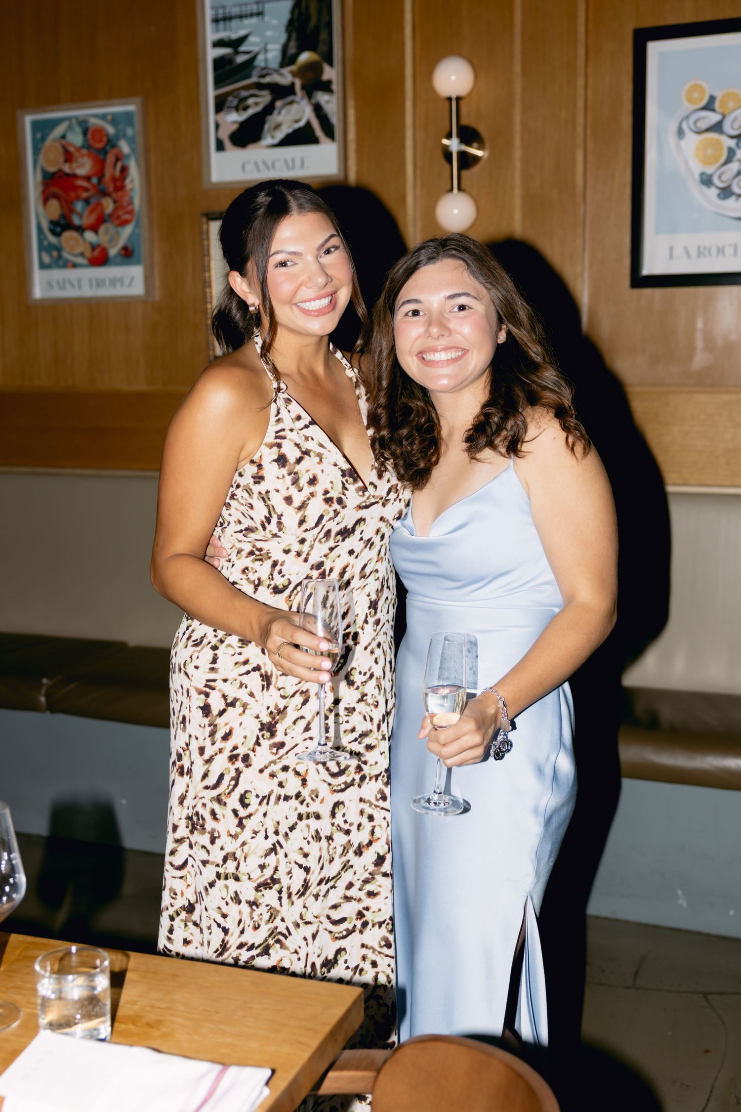 Two smiling women in elegant dresses holding champagne glasses pose together at a wedding dinner event.