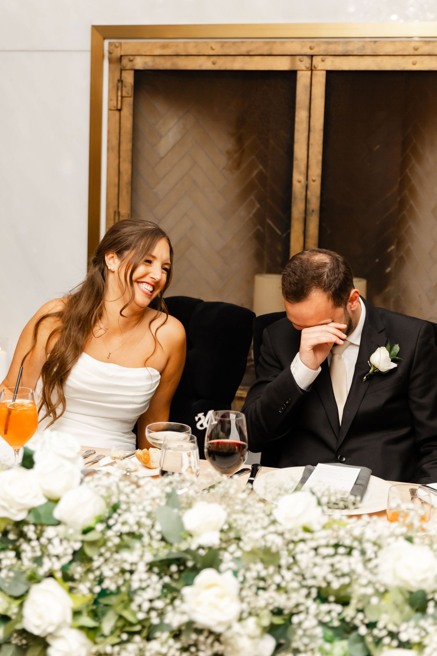 Bride and groom laugh together at sweetheart table adorned with white roses and greenery during reception.