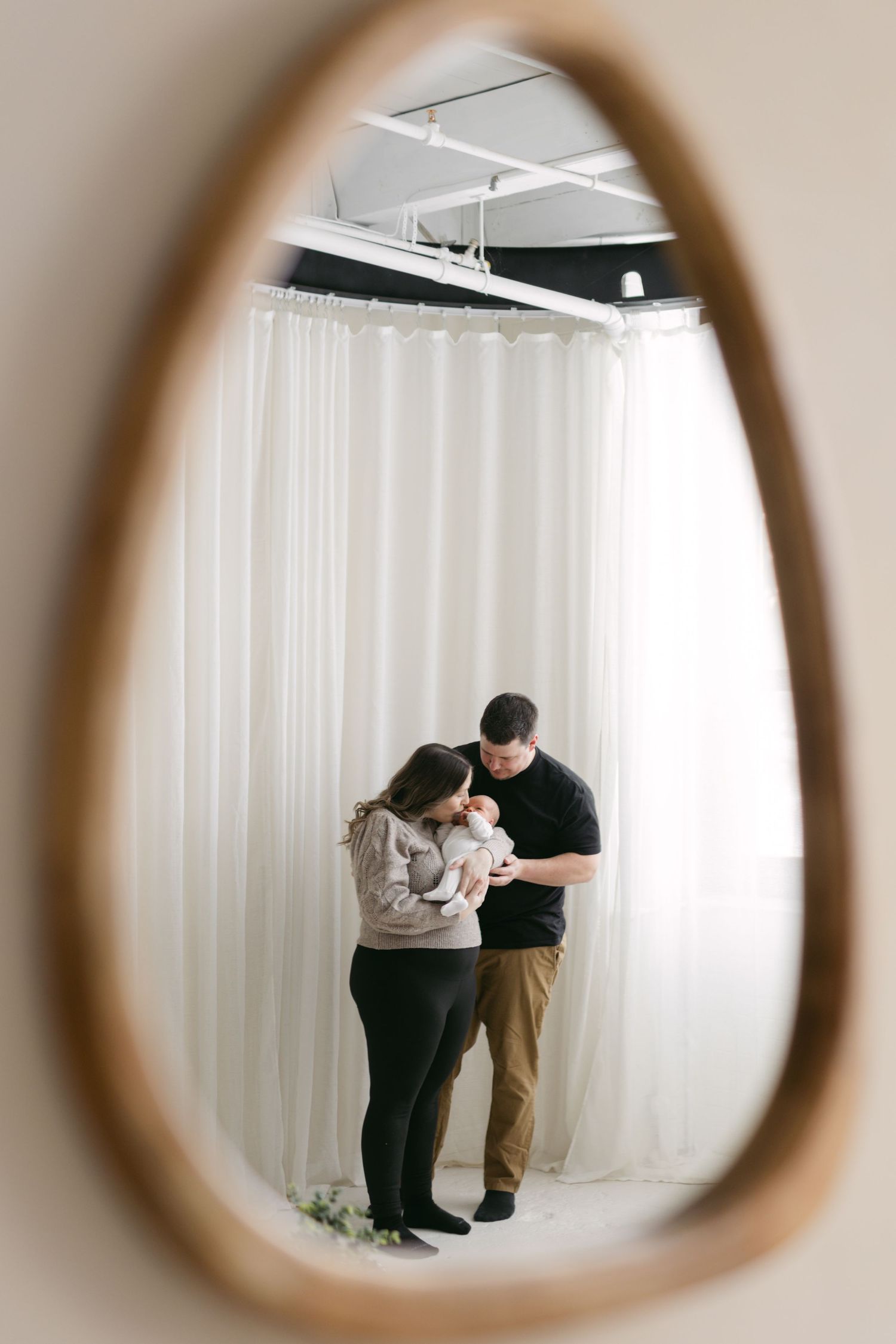 A couple stands together looking down at something in their hands, viewed through an oval mirror frame against white curtains.